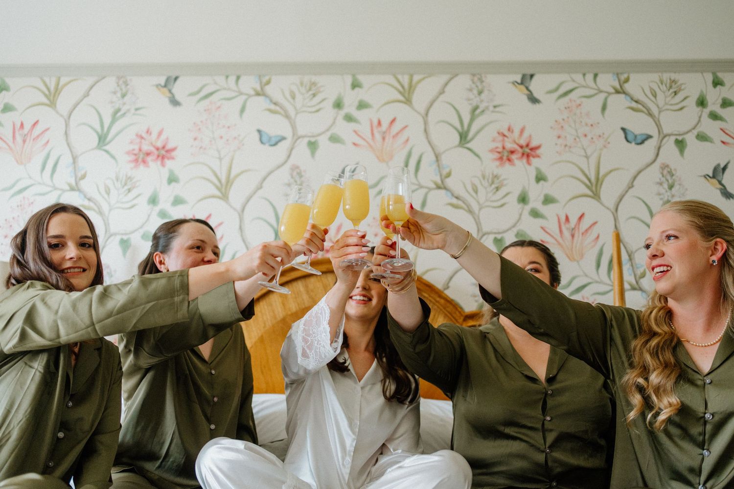Bridesmaids in green satin pajamas toasting with champagne glasses on a bed with floral wallpaper.