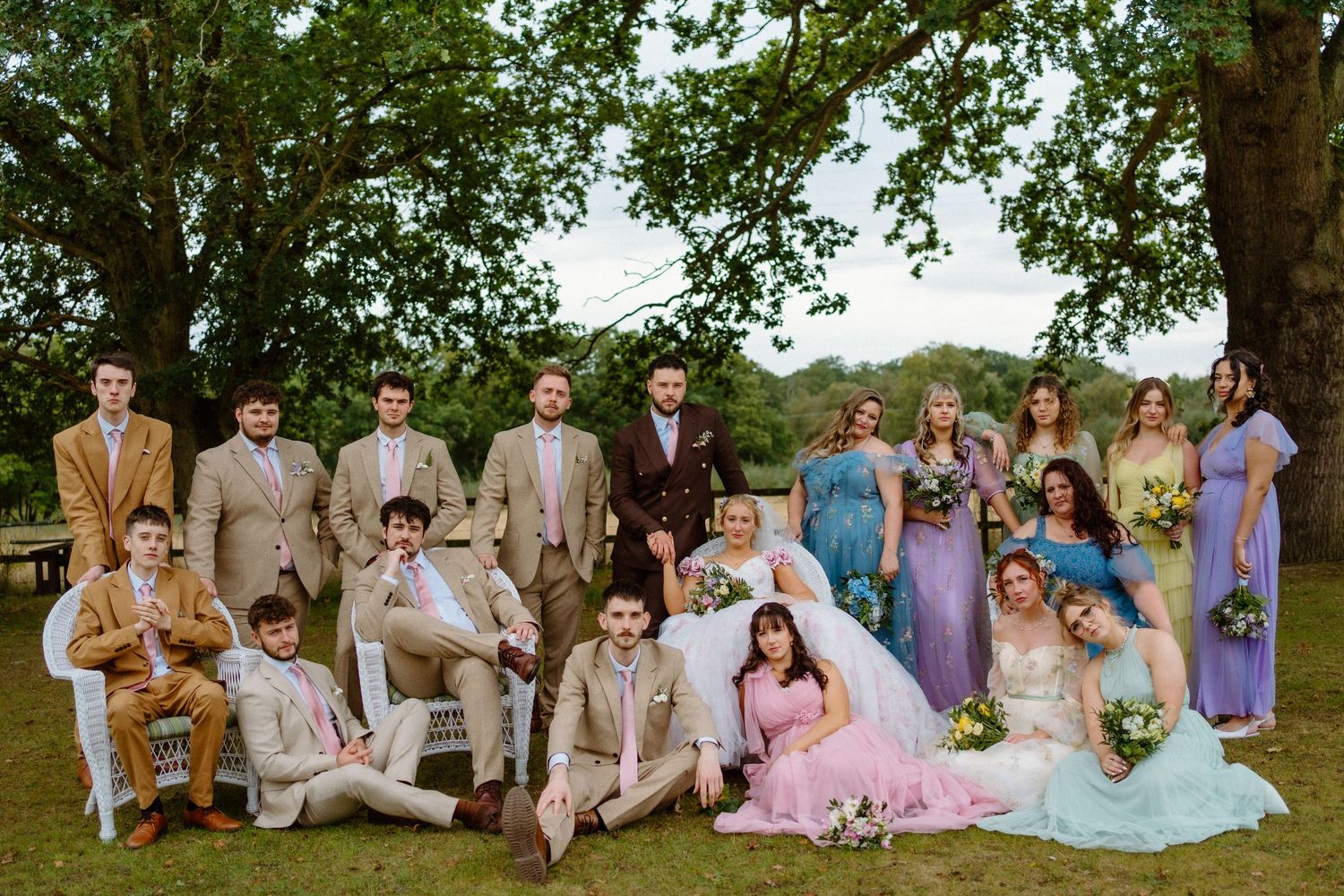 Large wedding party group portrait outdoors under tree with colorful bridesmaid dresses and tan groomsmen suits.