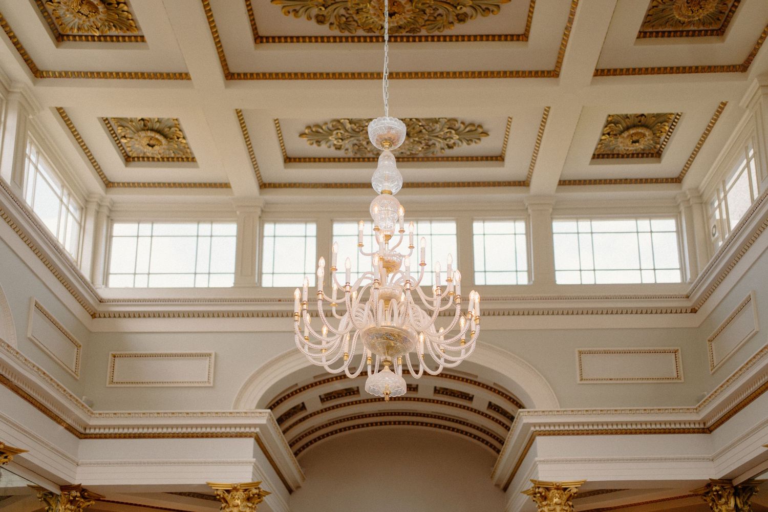 Stunning crystal chandelier hanging from an ornate gold and white coffered ceiling in an elegant wedding venue ballroom.
