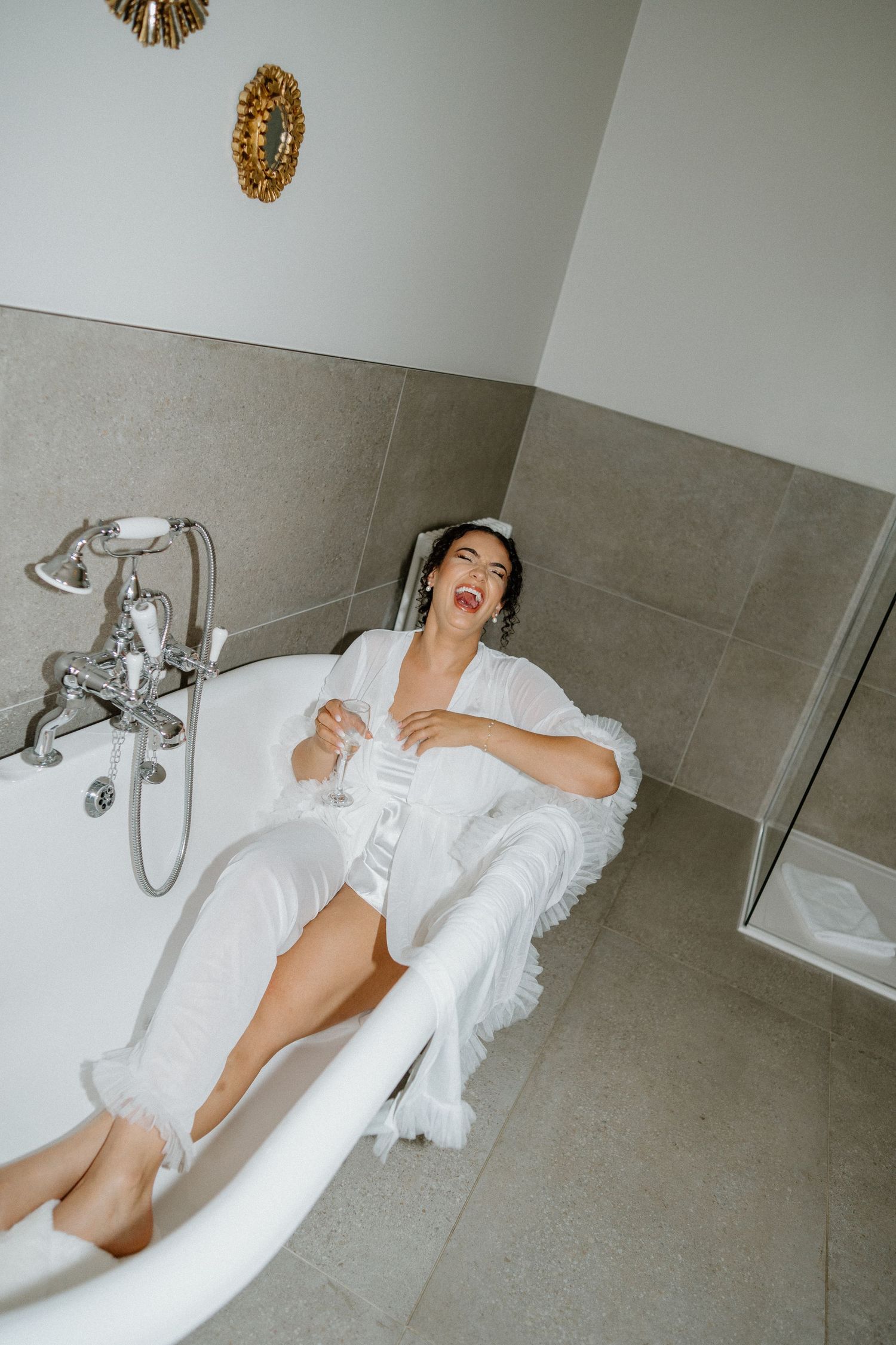 Bride in white robe relaxing in a clawfoot bathtub with champagne glass, laughing joyfully.