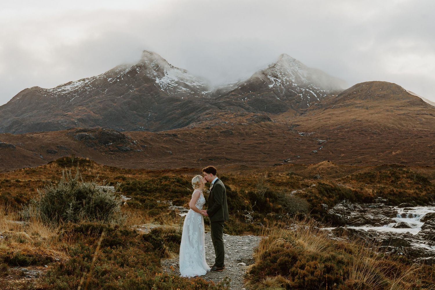 Bride and groom embrace on Scottish highland landscape with snow-capped mountains and dramatic cloudy sky.