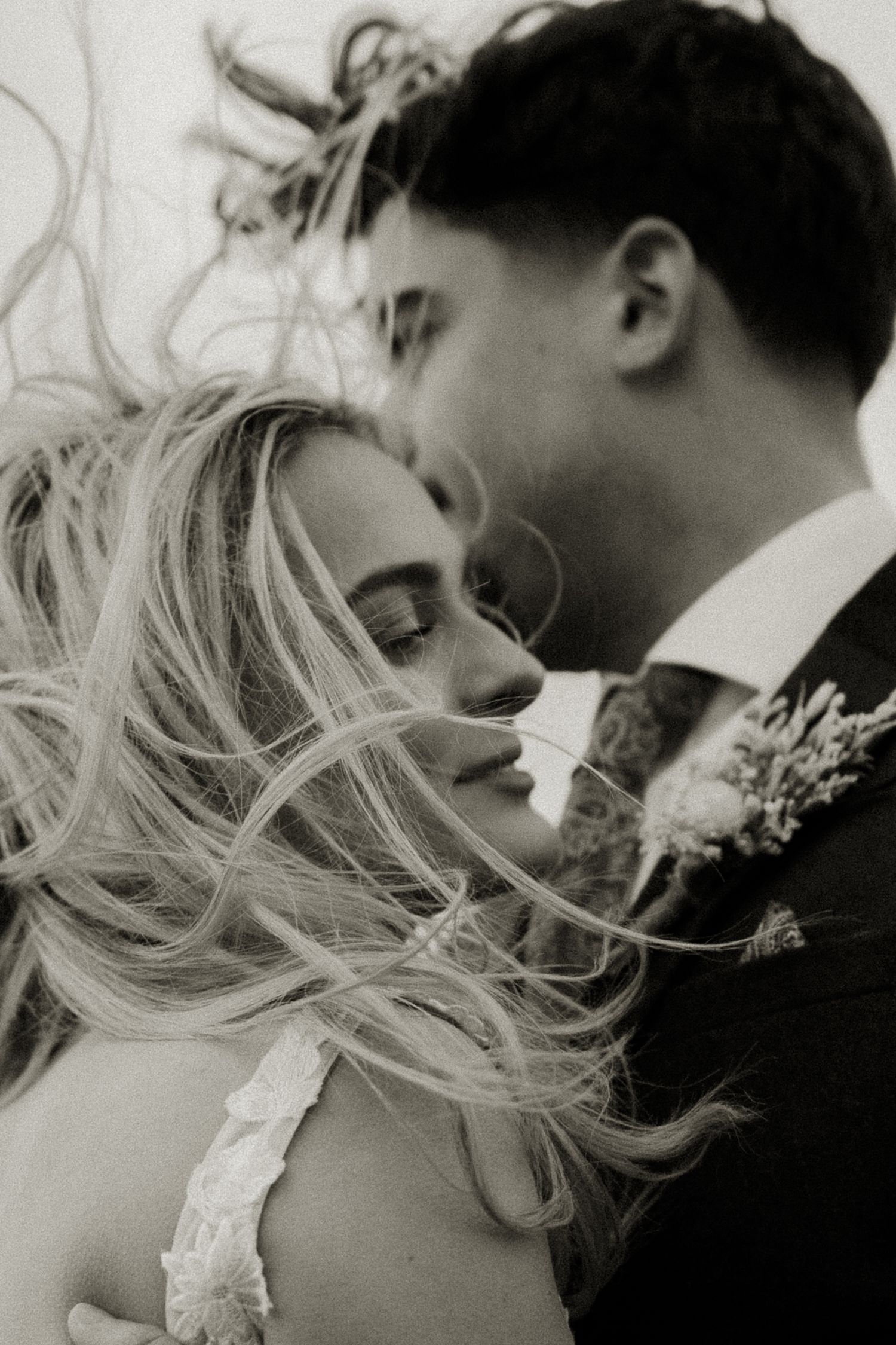 Black and white photo of bride with wind-blown hair and groom embracing at outdoor wedding.