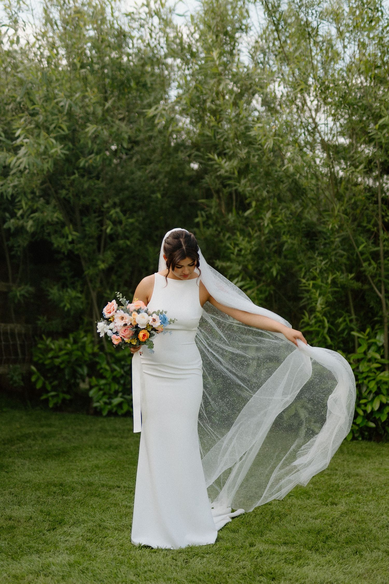Bride in white gown holding colorful bouquet while flowing veil catches the breeze in a lush garden.