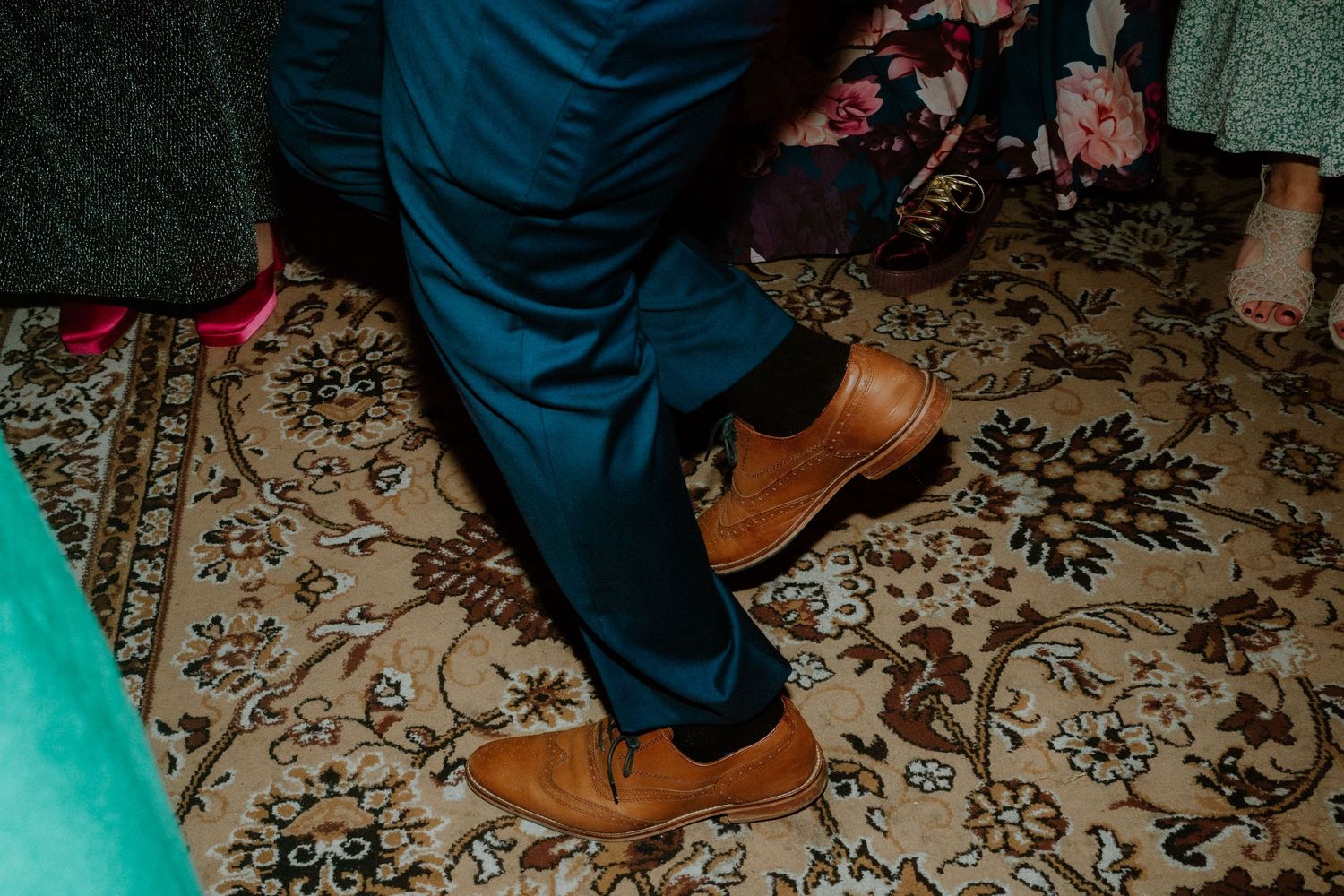 Close-up of wedding guest dancing in tan leather shoes on a patterned carpet.