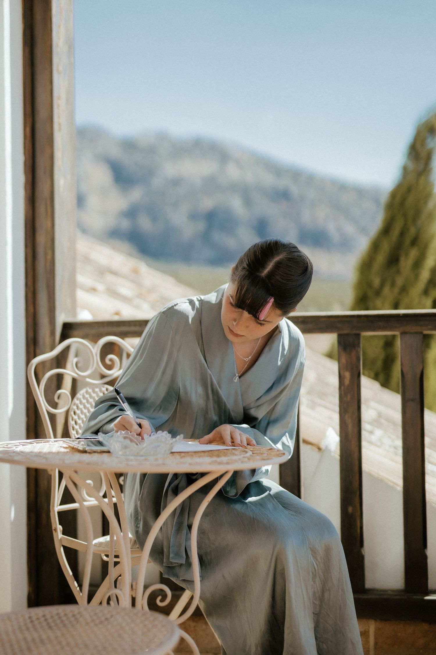 Bride in blue robe writes vows at ornate table on mountain balcony in sunlight.