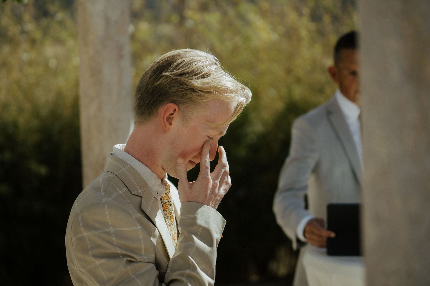 Emotional wedding guest in a plaid suit wipes tears during an outdoor wedding ceremony in bright sunlight.