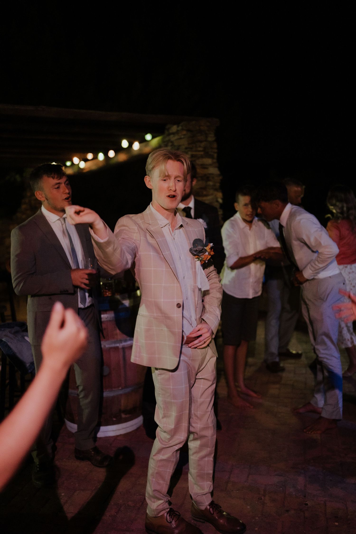 Blonde groom in plaid suit holding microphone while speaking at outdoor evening wedding reception.