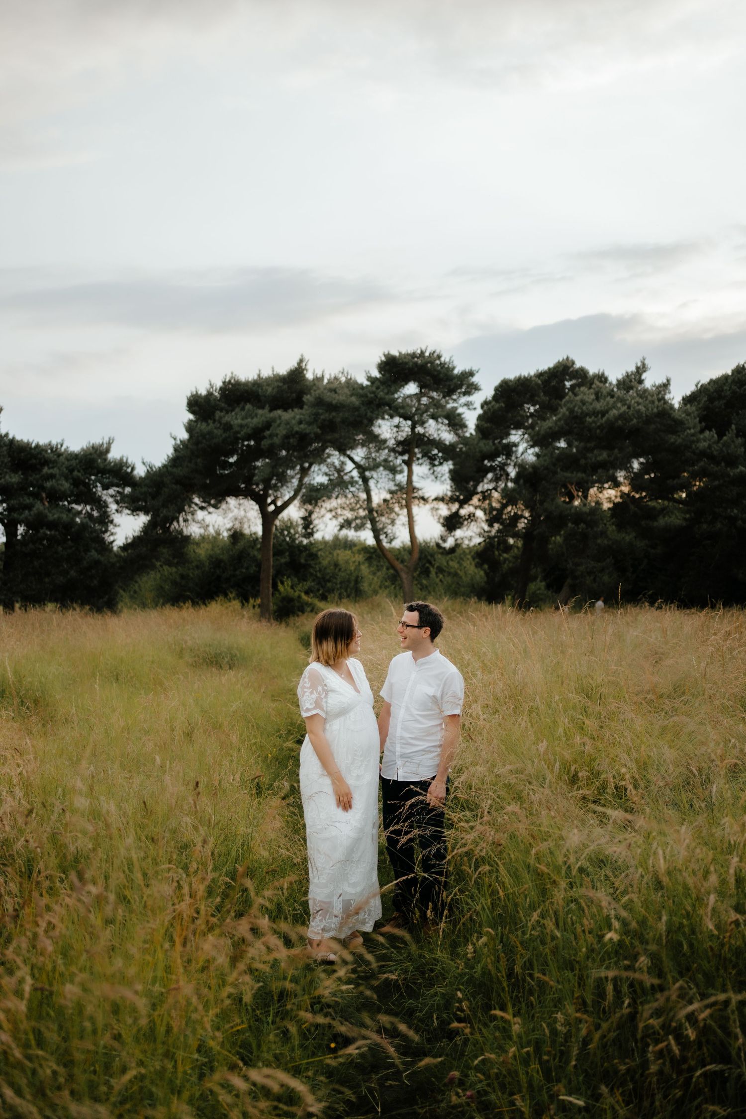 A pregnant couple holds hands while standing in a lush meadow at dusk.