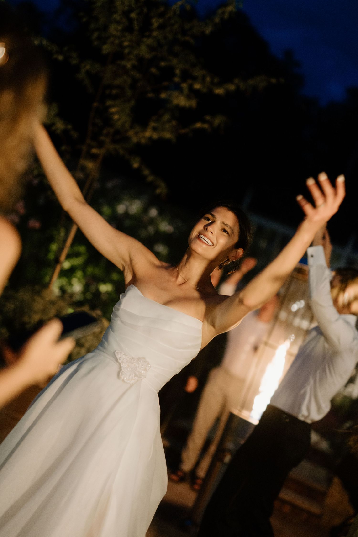 Bride in white strapless gown dancing joyfully with arms raised at outdoor evening wedding reception.