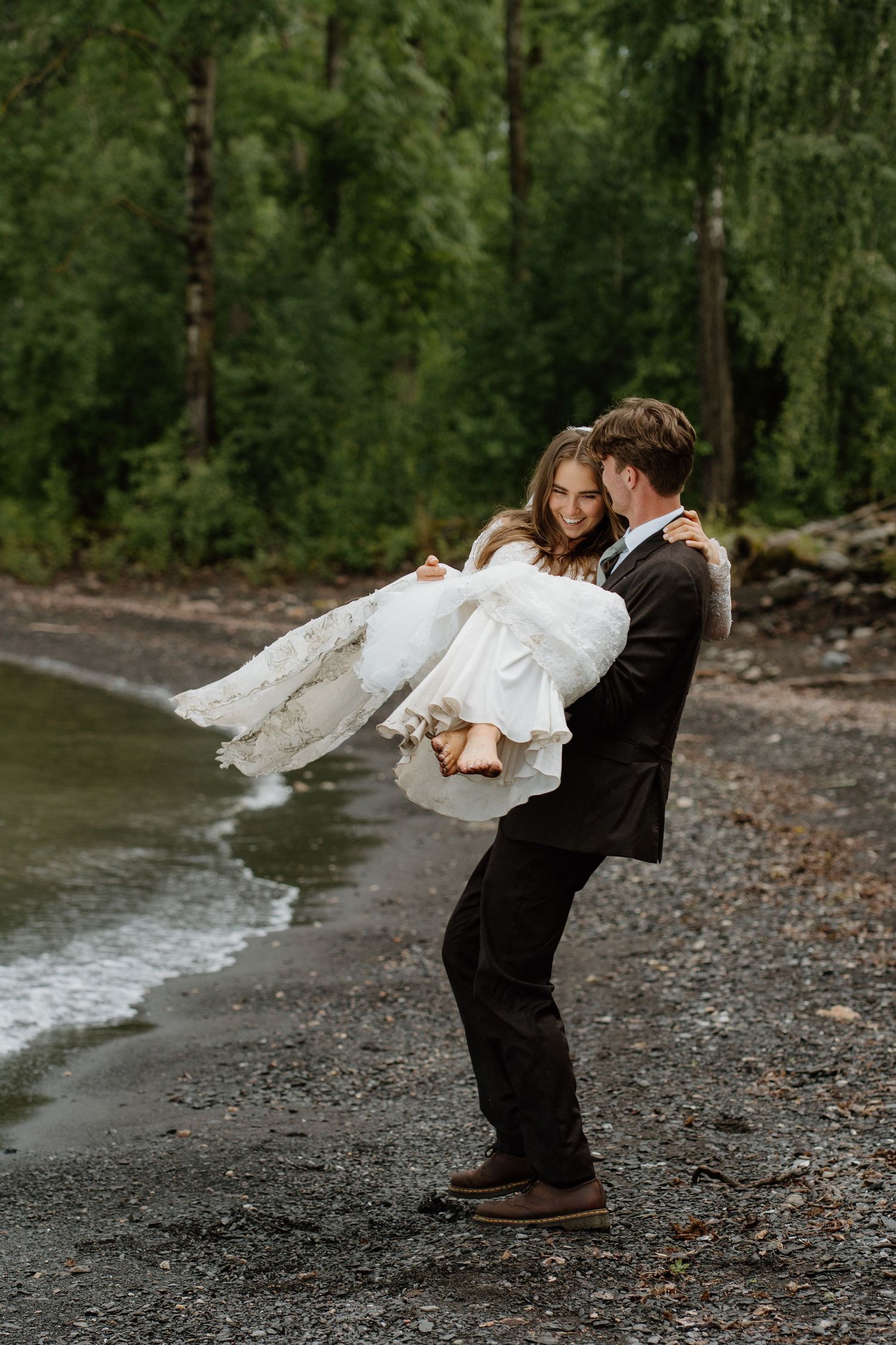 Groom carries bride in white lace dress along rocky beach with lush green forest backdrop.