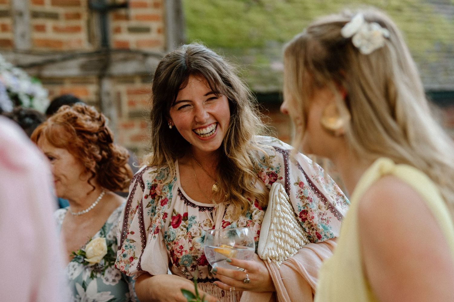 Joyful wedding guest in floral dress laughs while socializing outdoors at a rustic wedding reception.