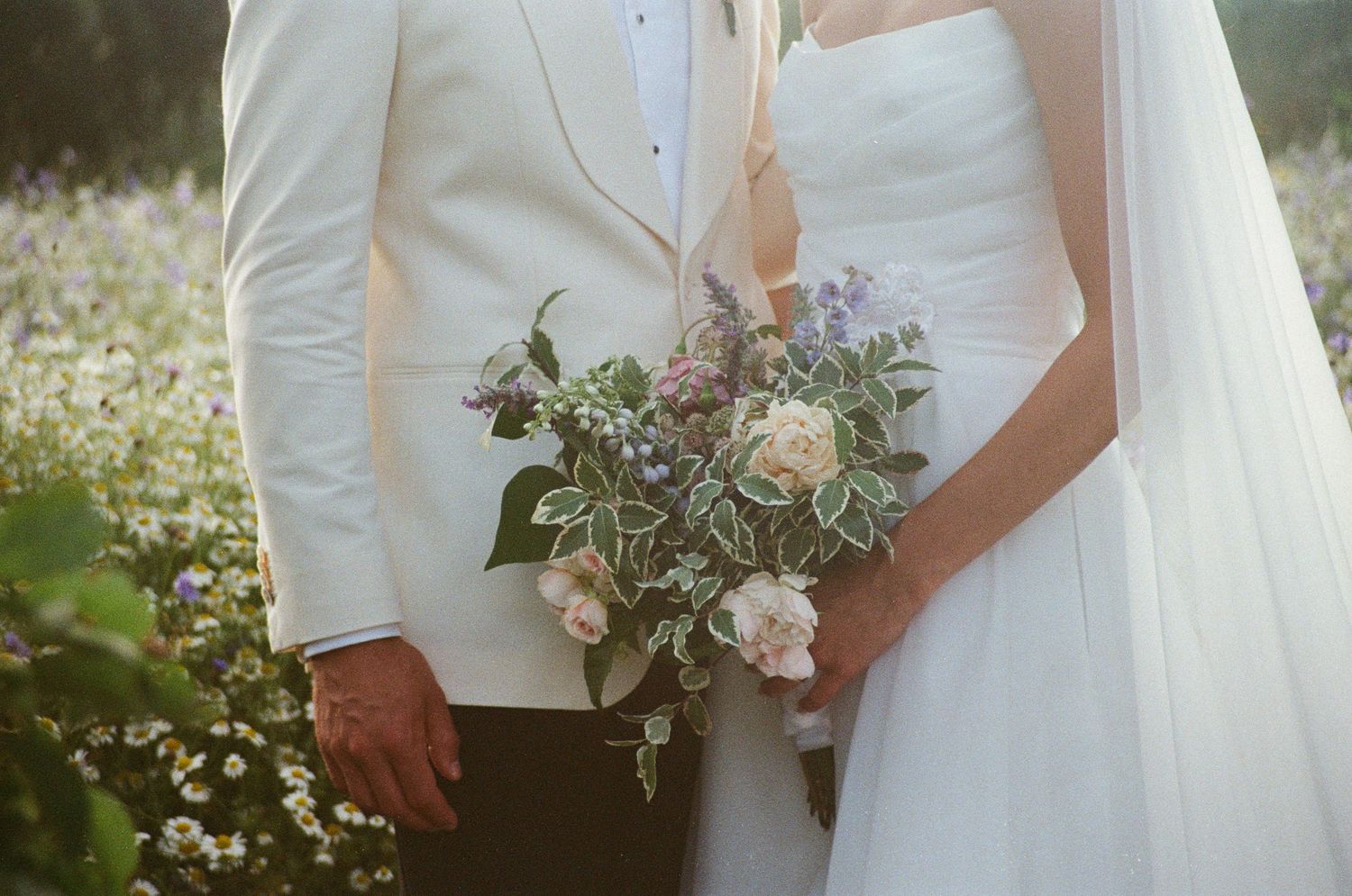 Close-up of bride and groom holding romantic wildflower bouquet in white and purple tones outdoors.