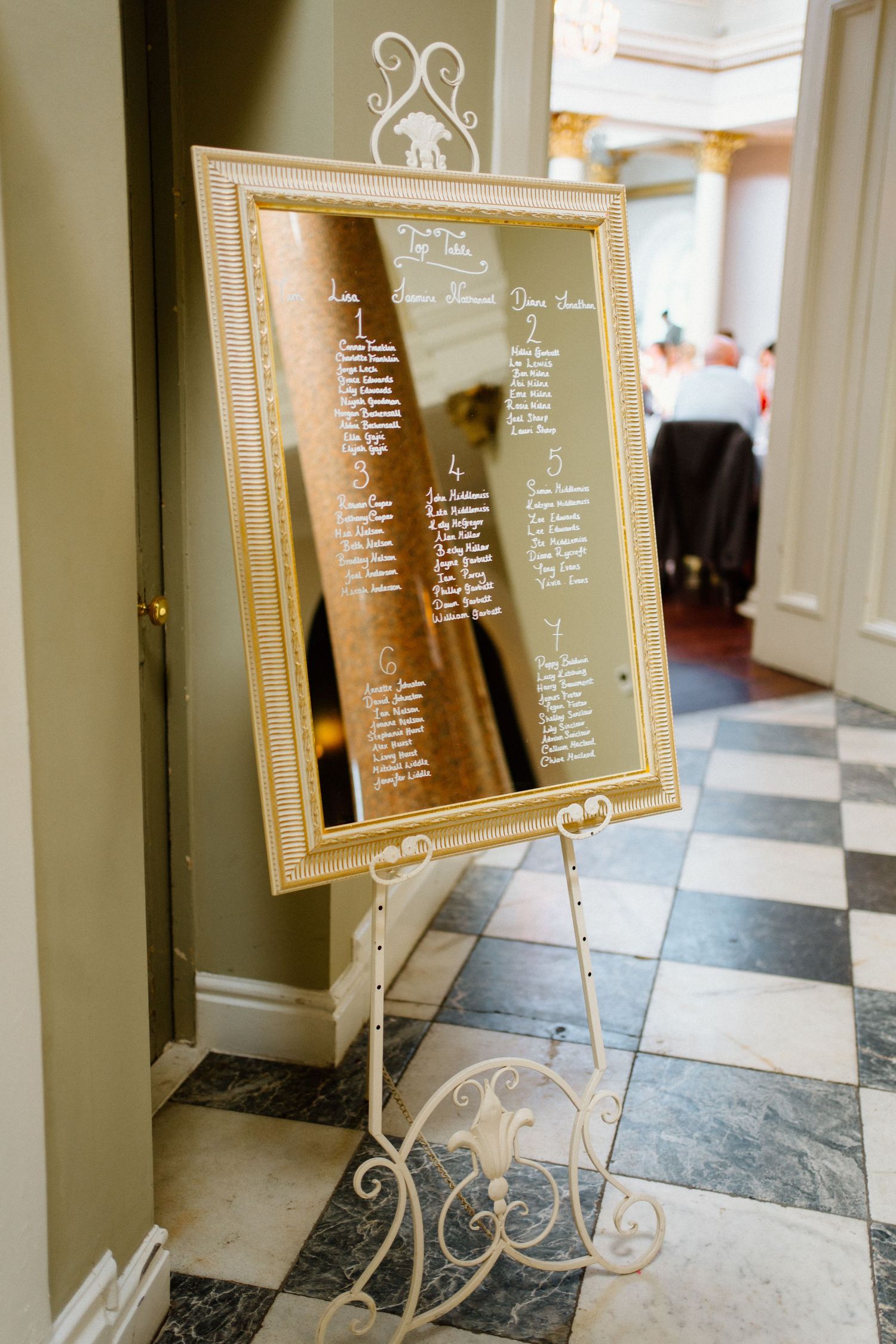 Elegant gold-framed mirror seating chart on a white ornate easel displayed on a checkered marble floor at a wedding.