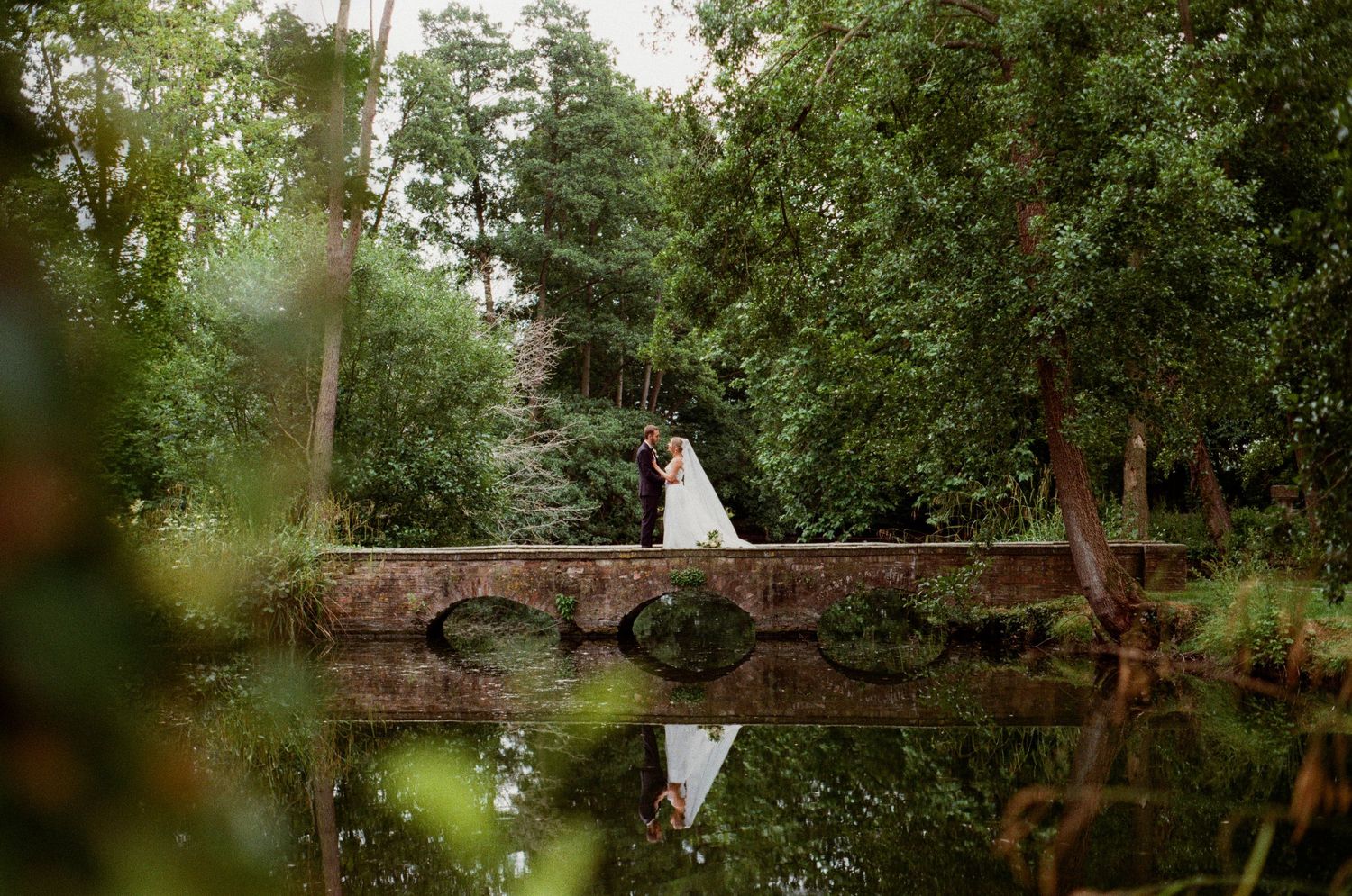Bride and groom kiss on a stone bridge reflected in calm water surrounded by lush green trees.