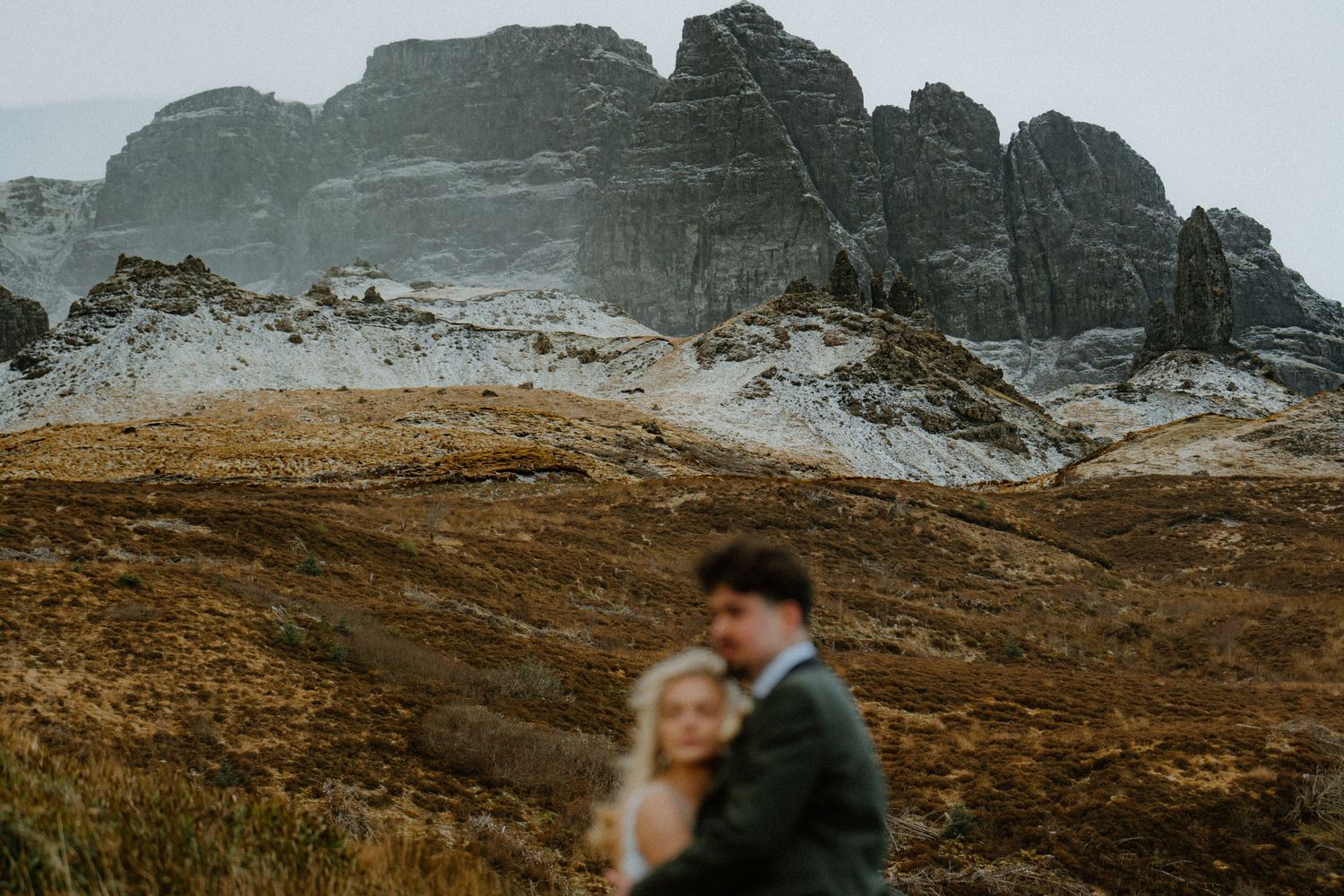 Wedding couple embraces in front of dramatic snow-dusted rocky Scottish Highland mountains.
