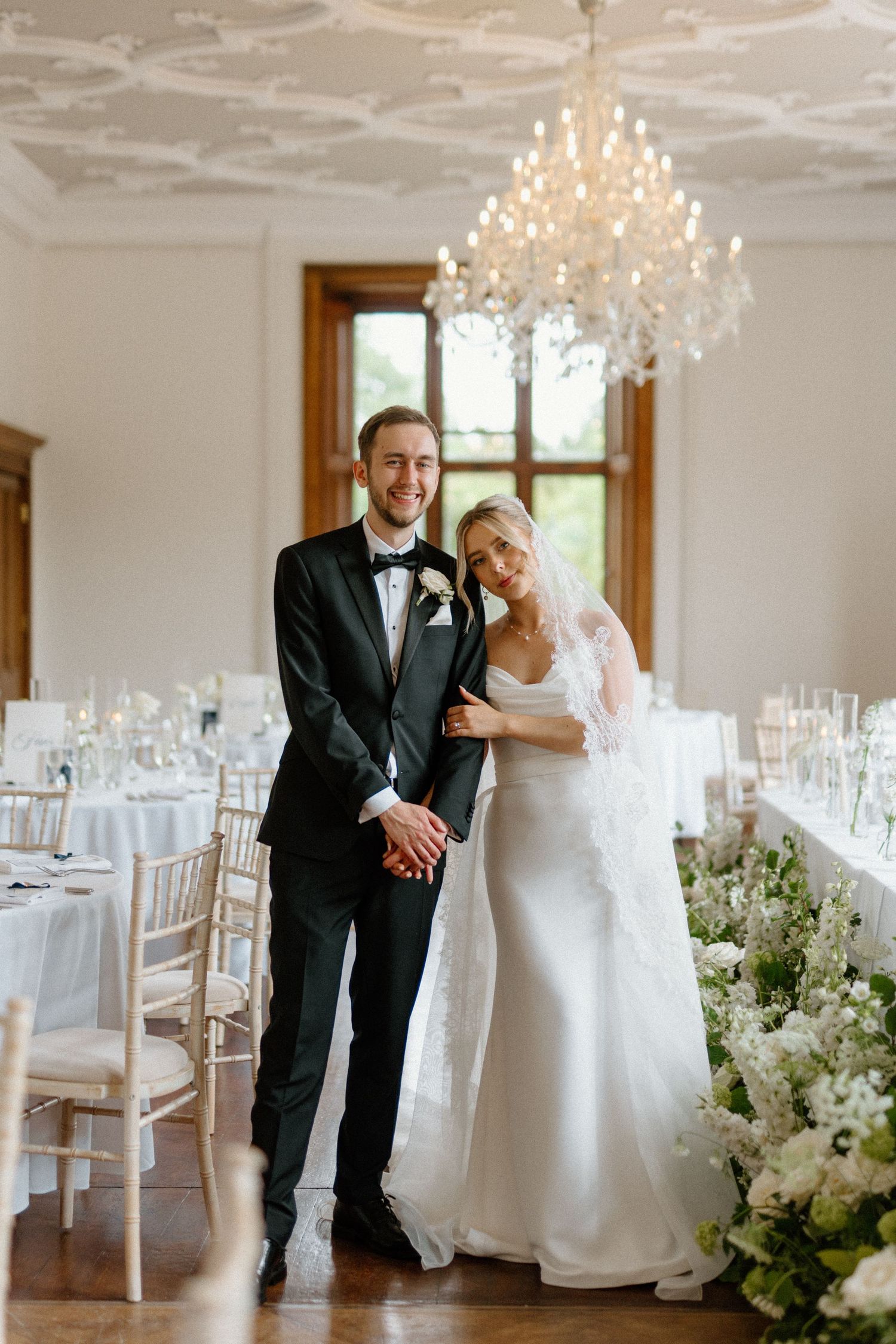 Smiling bride and groom posing in an elegant white reception room with crystal chandelier and floral decor.