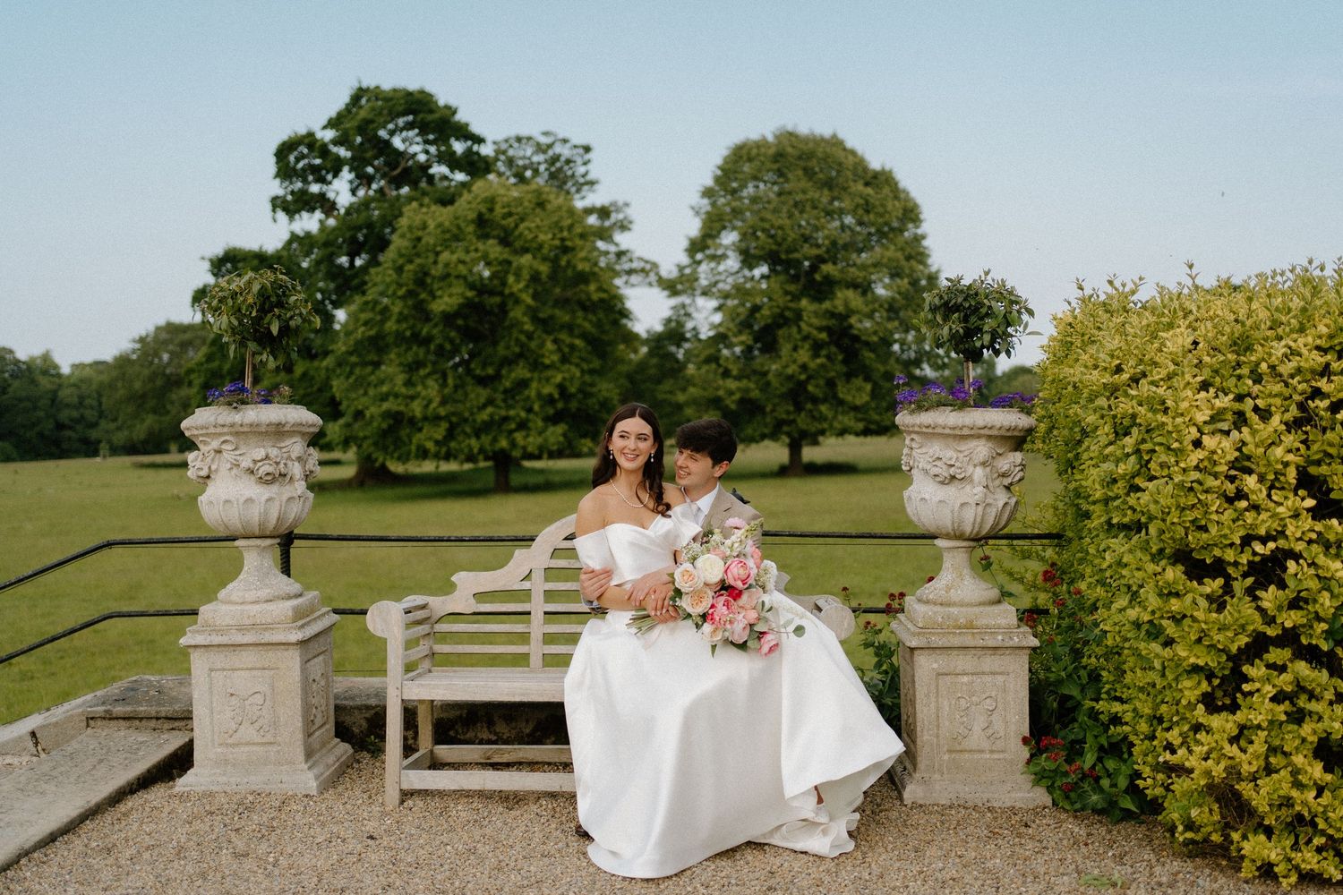 Two brides sitting together on a garden bench with a pink bouquet in sunny grounds.