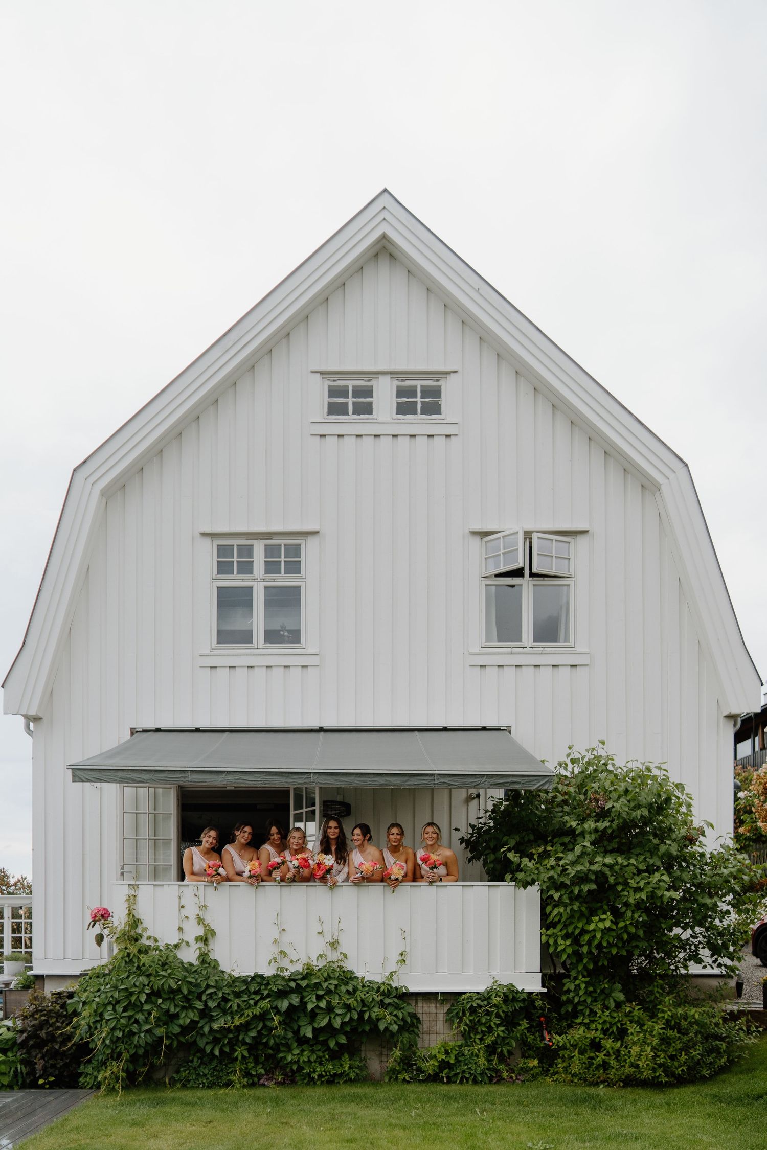 Bridesmaids in coral dresses stand on balcony of white Scandinavian farmhouse wedding venue.
