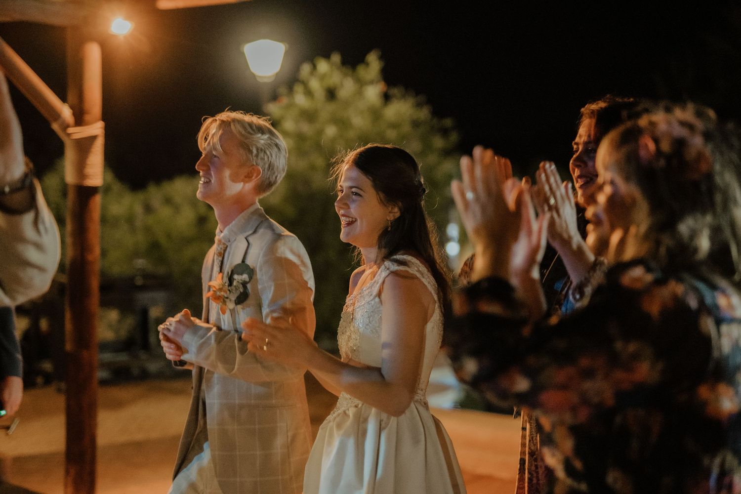Bride and groom laughing joyfully with guests clapping at outdoor nighttime wedding reception.