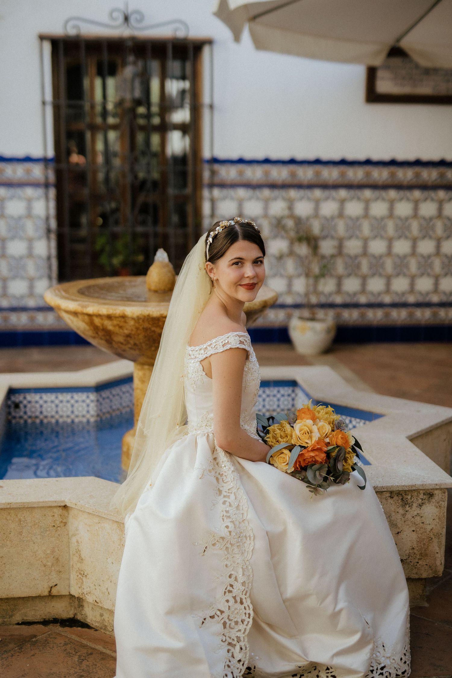 Smiling bride in off-shoulder white lace gown sits by a tiled fountain holding a yellow and orange bouquet.