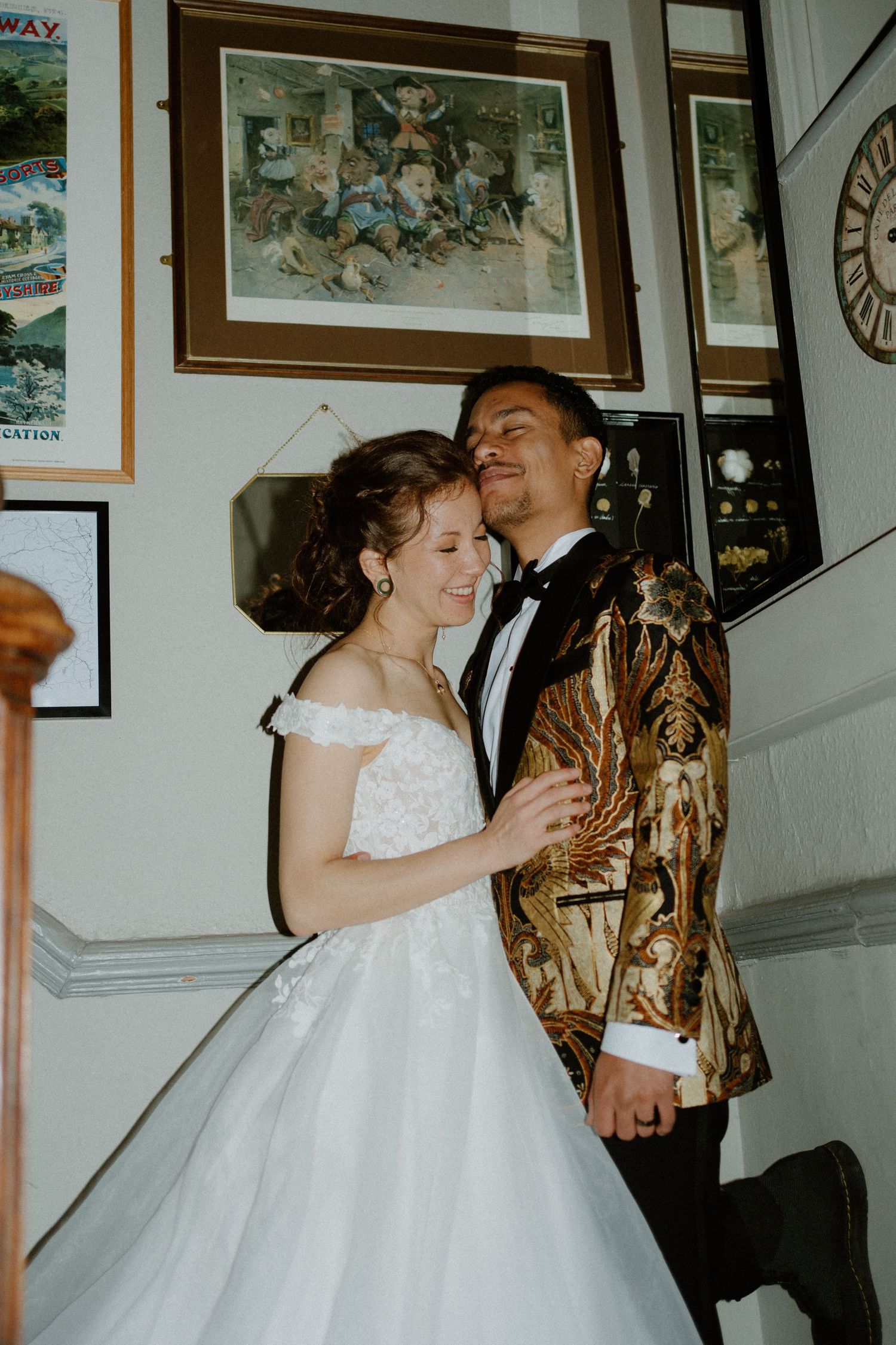 Bride and groom share a joyful moment on staircase, groom wearing ornate gold patterned blazer with black tuxedo.
