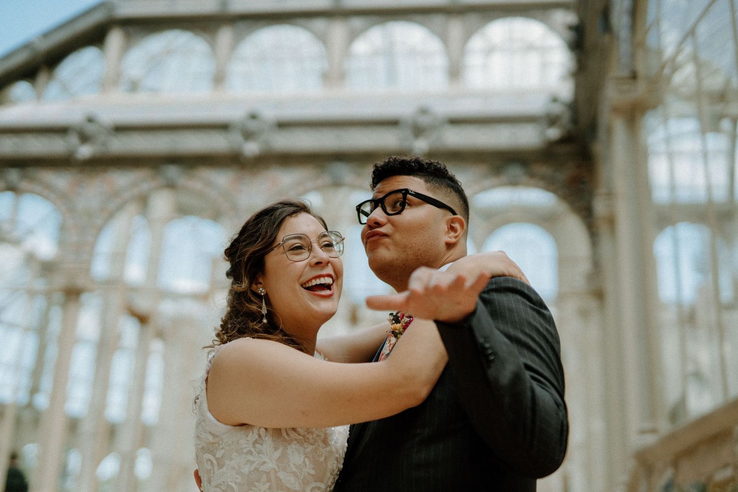 Laughing bride and groom in matching glasses dance together outside ornate glass building.