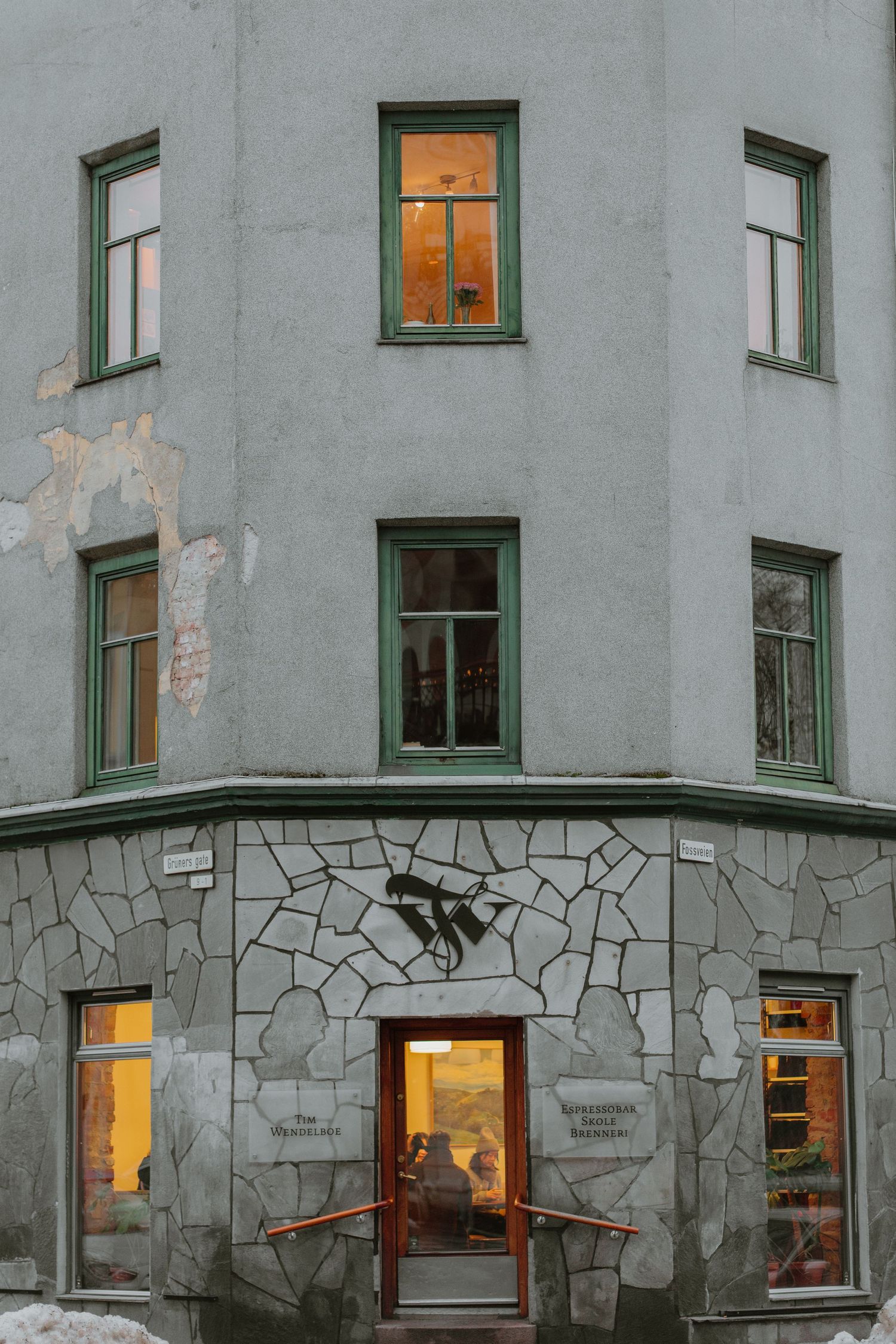 Corner view of a grey apartment building with green-trimmed windows and stone base featuring warm glowing lights at dusk.