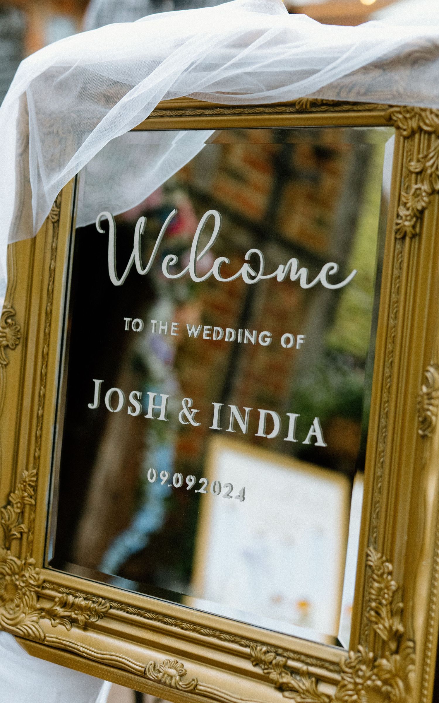 A gold-framed mirror wedding welcome sign with white text displays the date 09.09.2024 with flowing white fabric draping.