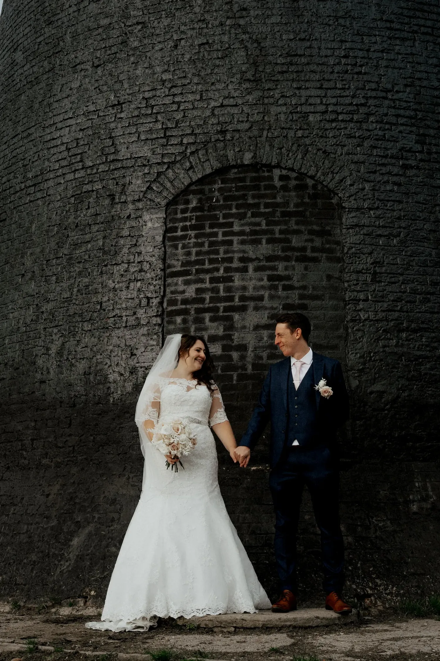 Newlyweds pose against dramatic brick wall architecture in formal wedding attire.