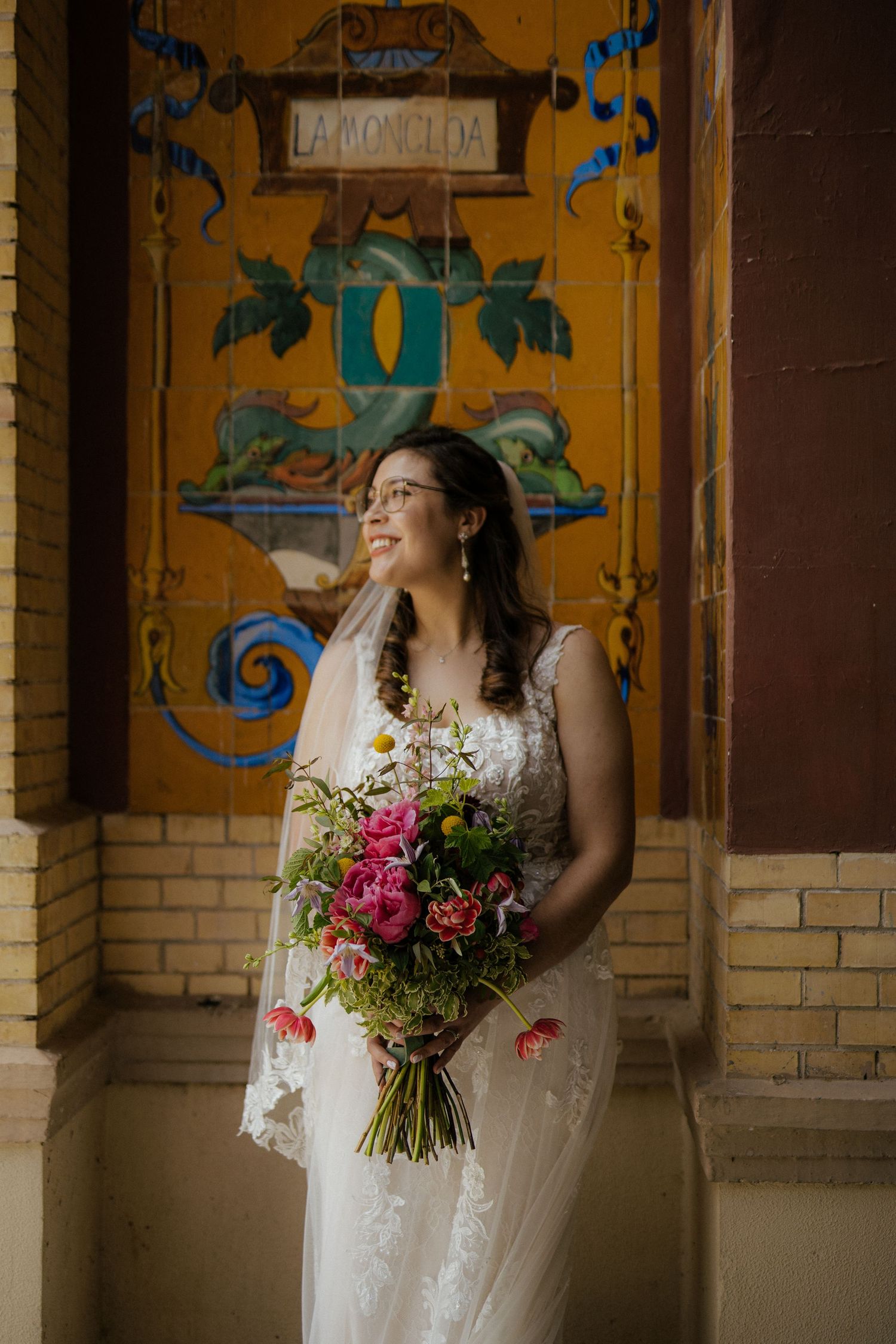 Smiling bride in lace gown holding a colorful bouquet standing before a vibrant painted mural wall.