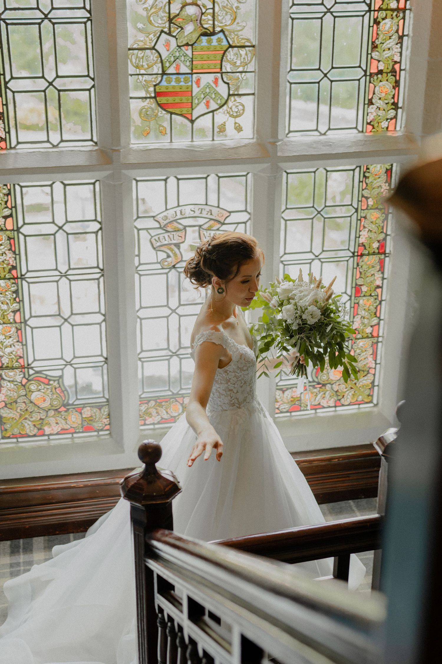 Bride in lace gown descends staircase before beautiful stained glass window holding bouquet.