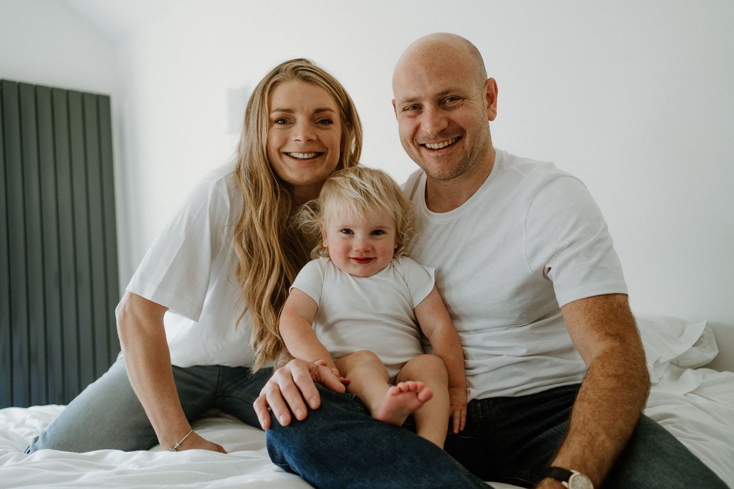 Happy family portrait of smiling parents with their baby sitting on a white bed.