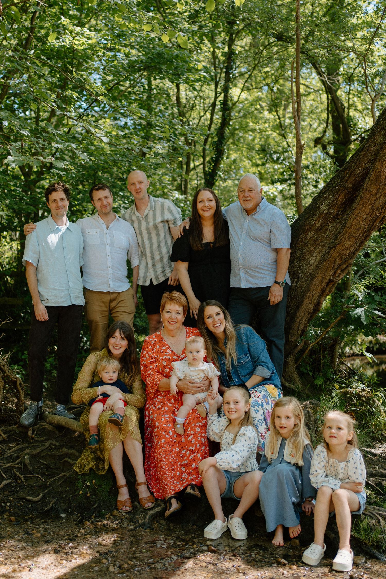 A large multigenerational family poses together near a large tree in a green woodland.