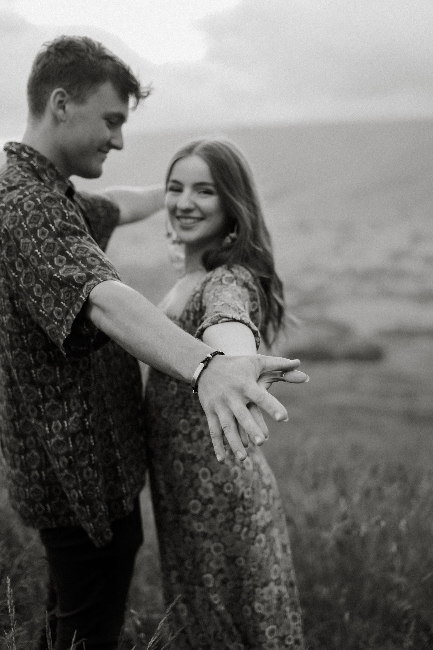 Black and white photo of smiling couple holding hands in a field, showcasing engagement ring.