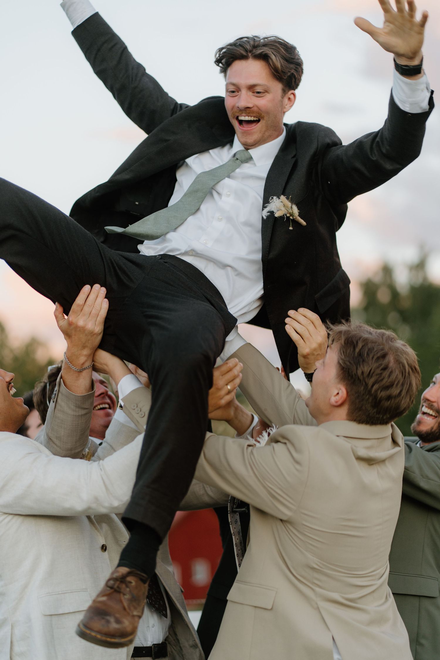 Joyful groom in black suit tossed into air by laughing groomsmen during outdoor wedding celebration.