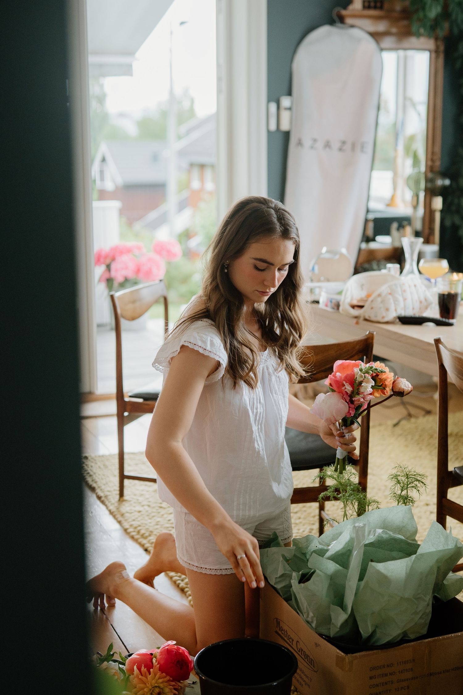 Bride in white dress arranging fresh flowers from a delivery box in a bright, cozy home interior.