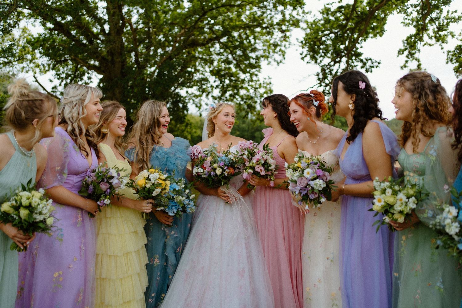 Bride in blush gown surrounded by bridesmaids in rainbow-colored dresses holding wildflower bouquets.