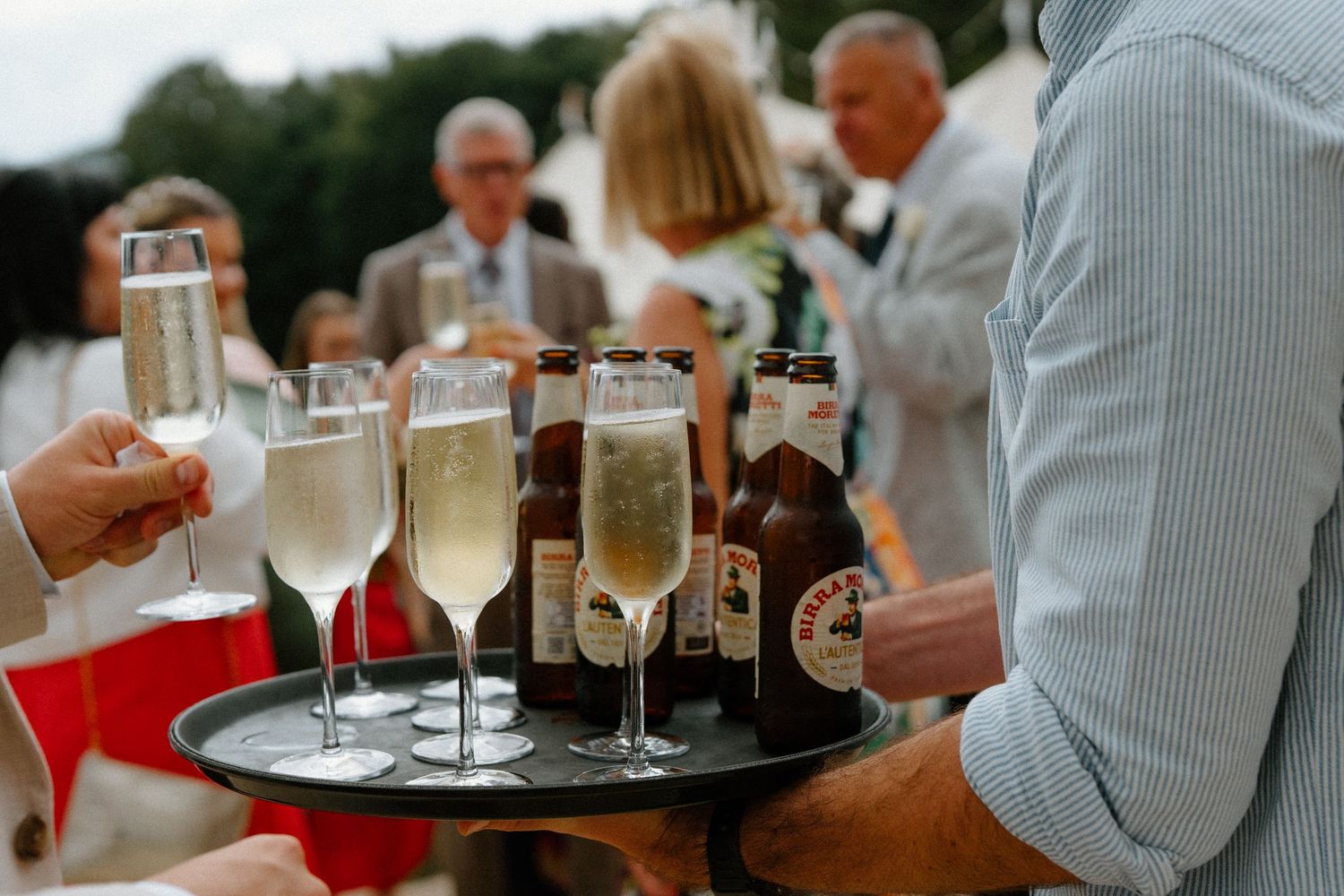 Wedding server carries tray of champagne flutes and beer bottles during outdoor cocktail reception.