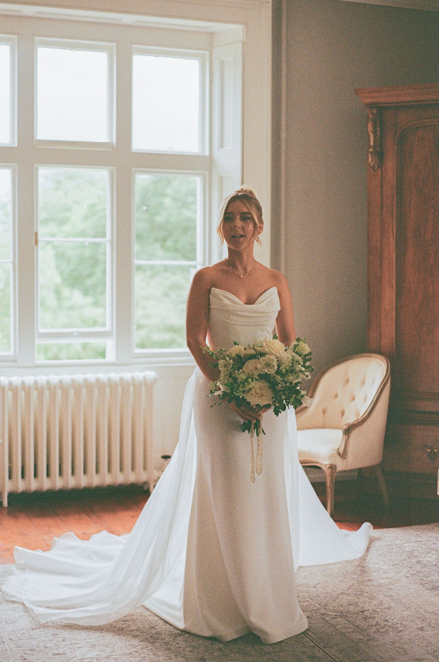 Elegant bride in strapless white gown holding green and white bouquet standing before large window in vintage room.