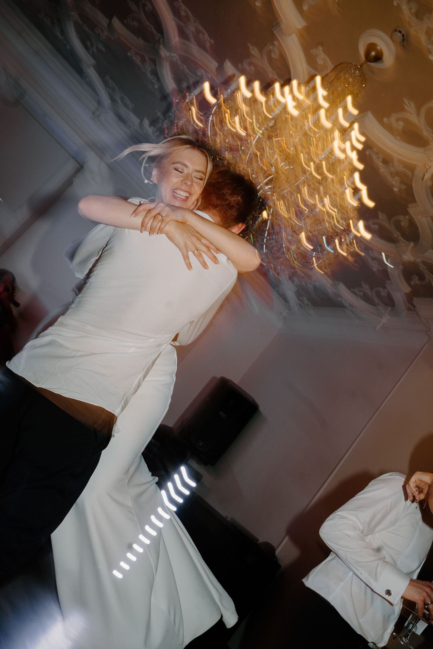 Groom lifts laughing bride during energetic wedding reception dance beneath ornate golden chandelier.
