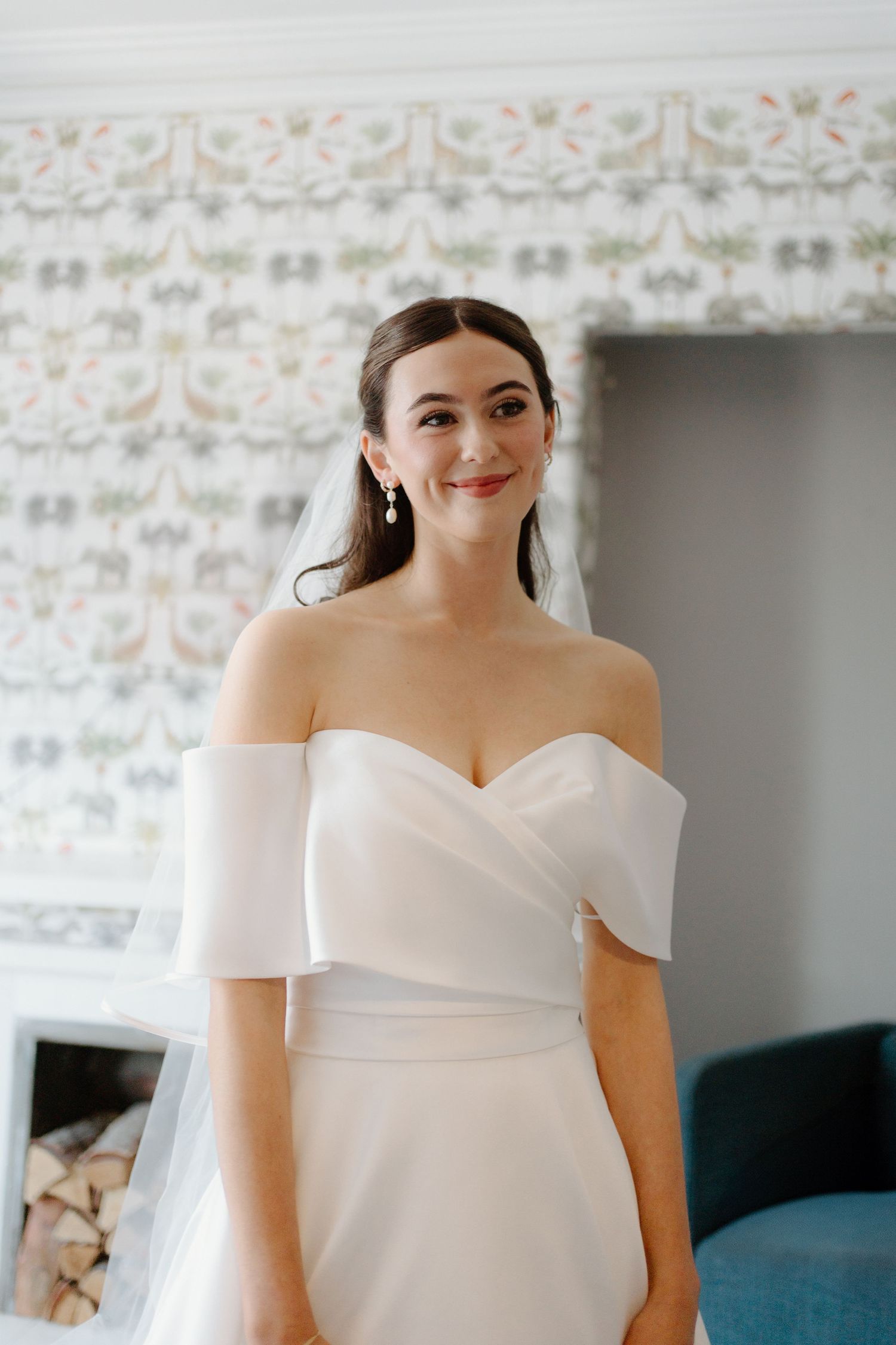 Elegant bride in off-shoulder white wedding gown with veil smiling in a decorated room.