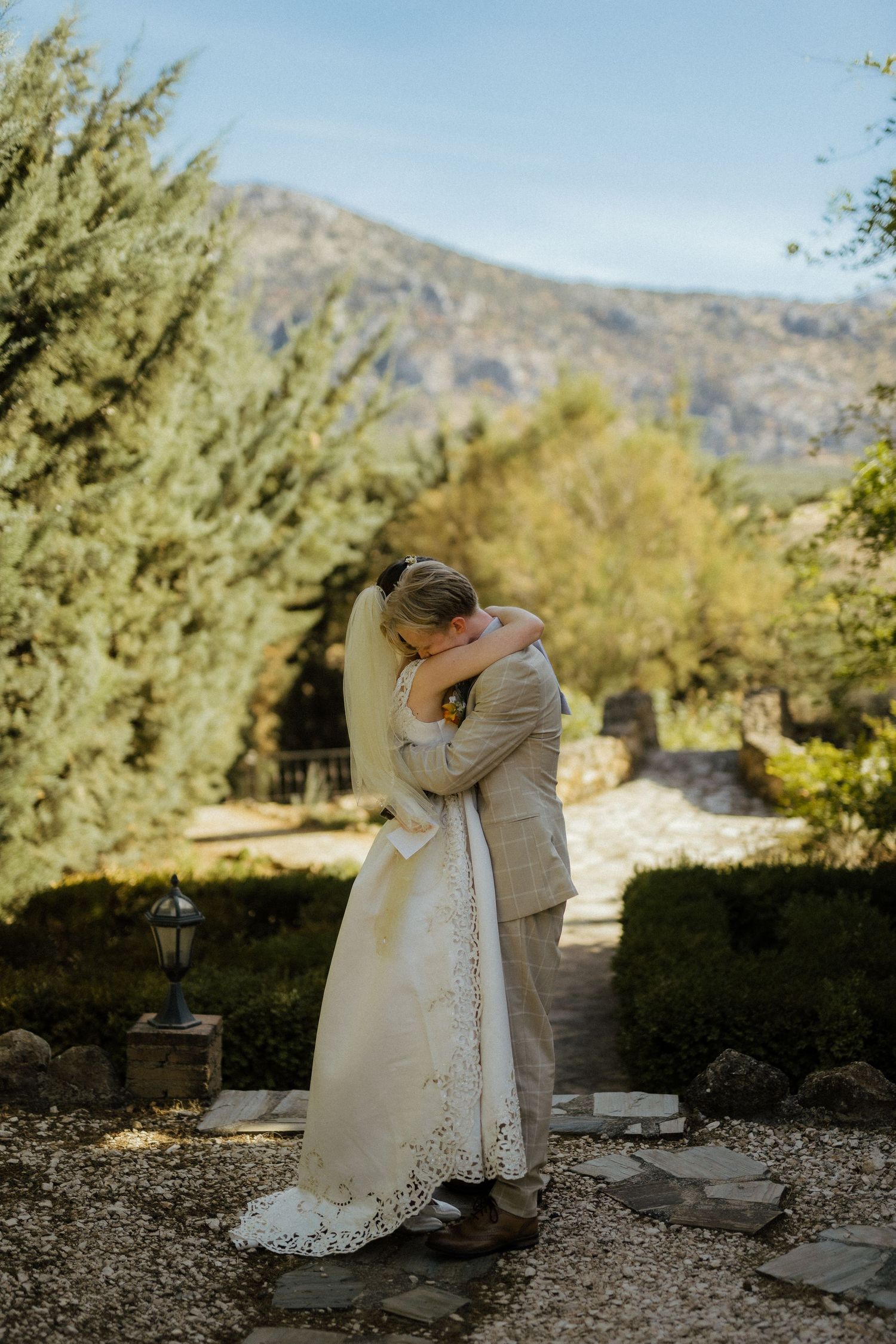 Bride and groom share emotional embrace on garden path with mountains behind.