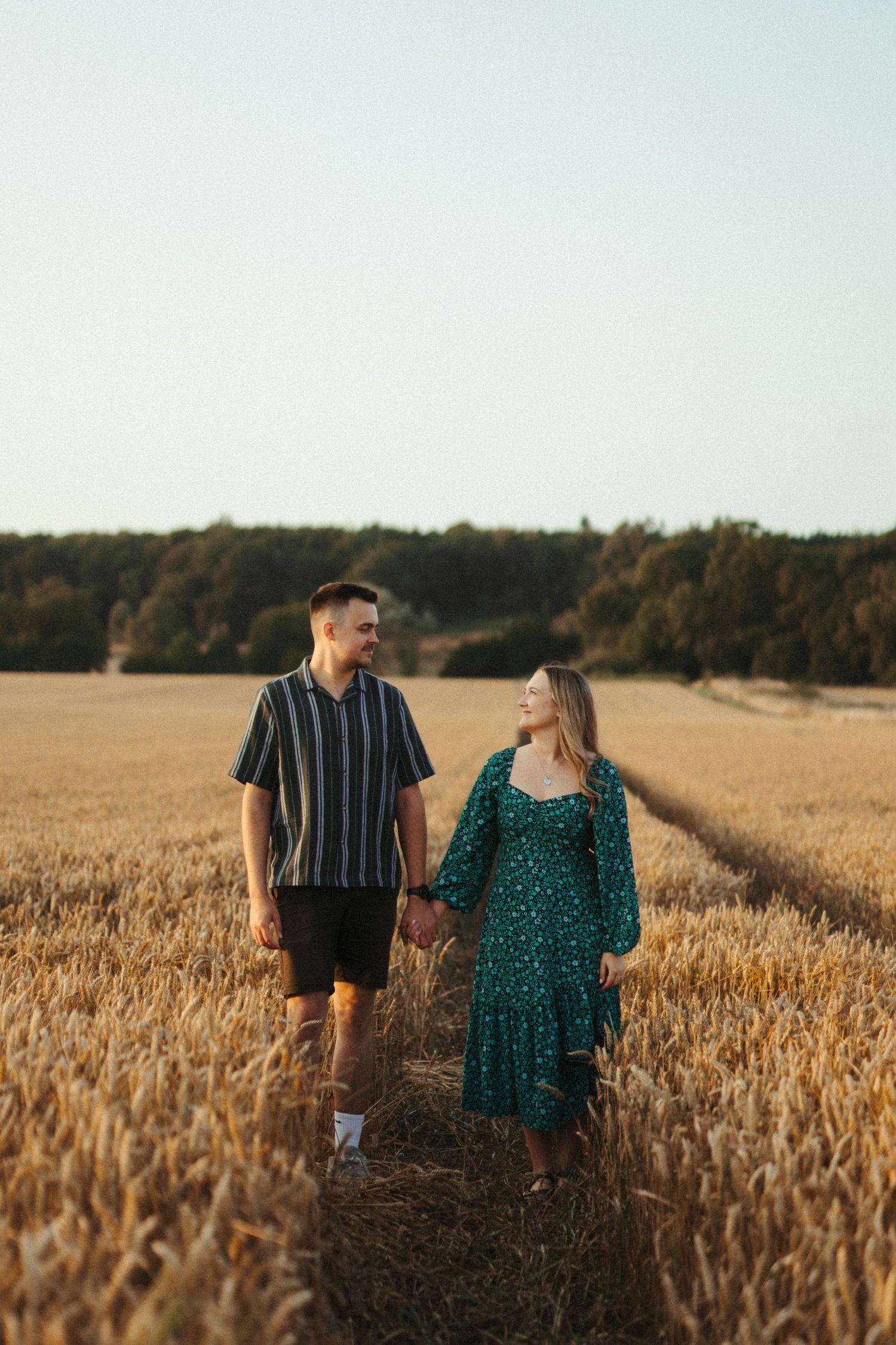 Couple walking hand in hand through a golden wheat field at sunset with forest in background.