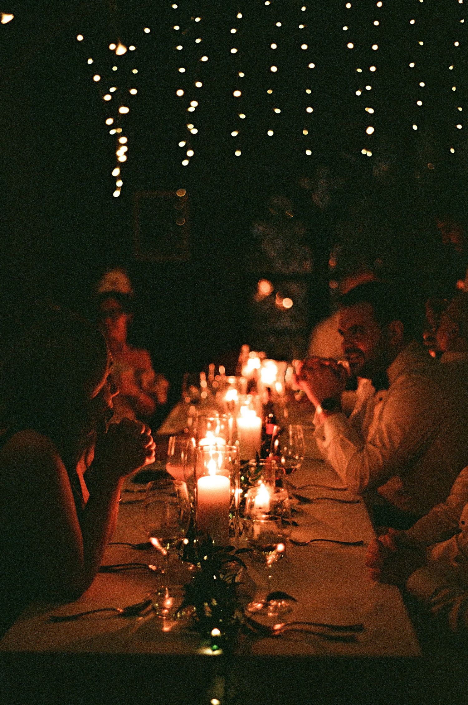 Wedding guests dining by candlelight at a long table under twinkling string lights at night.