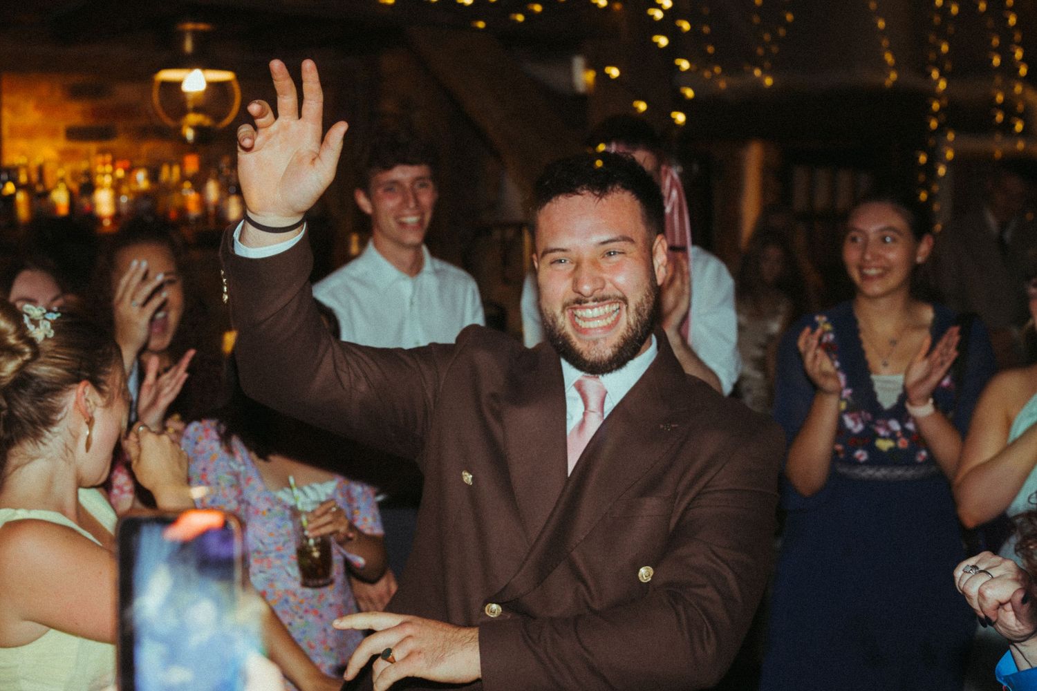 Smiling groom in brown suit waves to crowd while guests clap and celebrate at wedding reception.