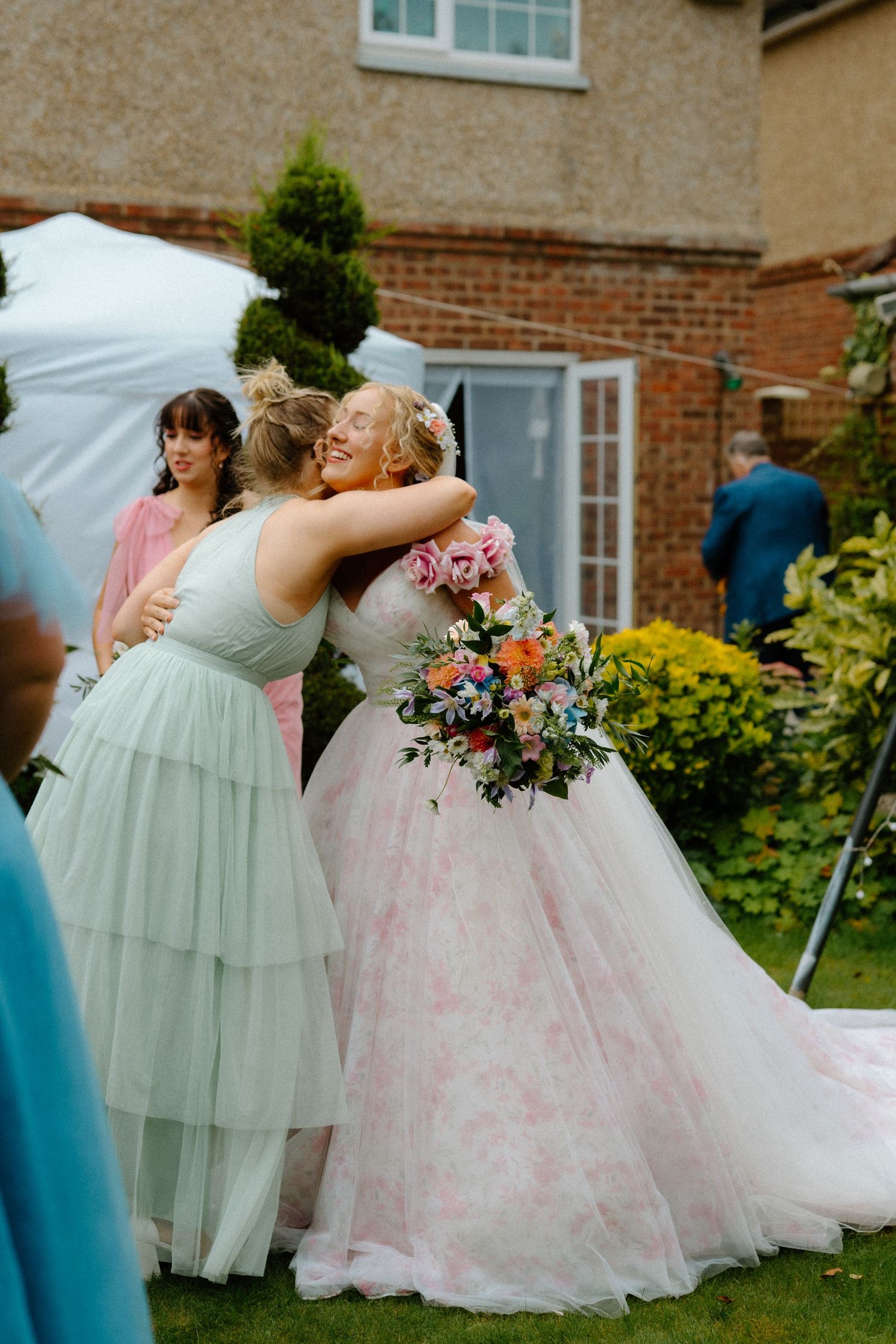 Bride in pink floral ball gown embracing a guest while holding colorful bouquet at outdoor wedding.