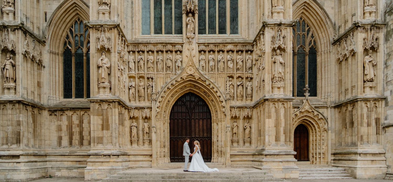 Bride and groom standing before grand ornate Gothic cathedral entrance in stunning wedding attire.