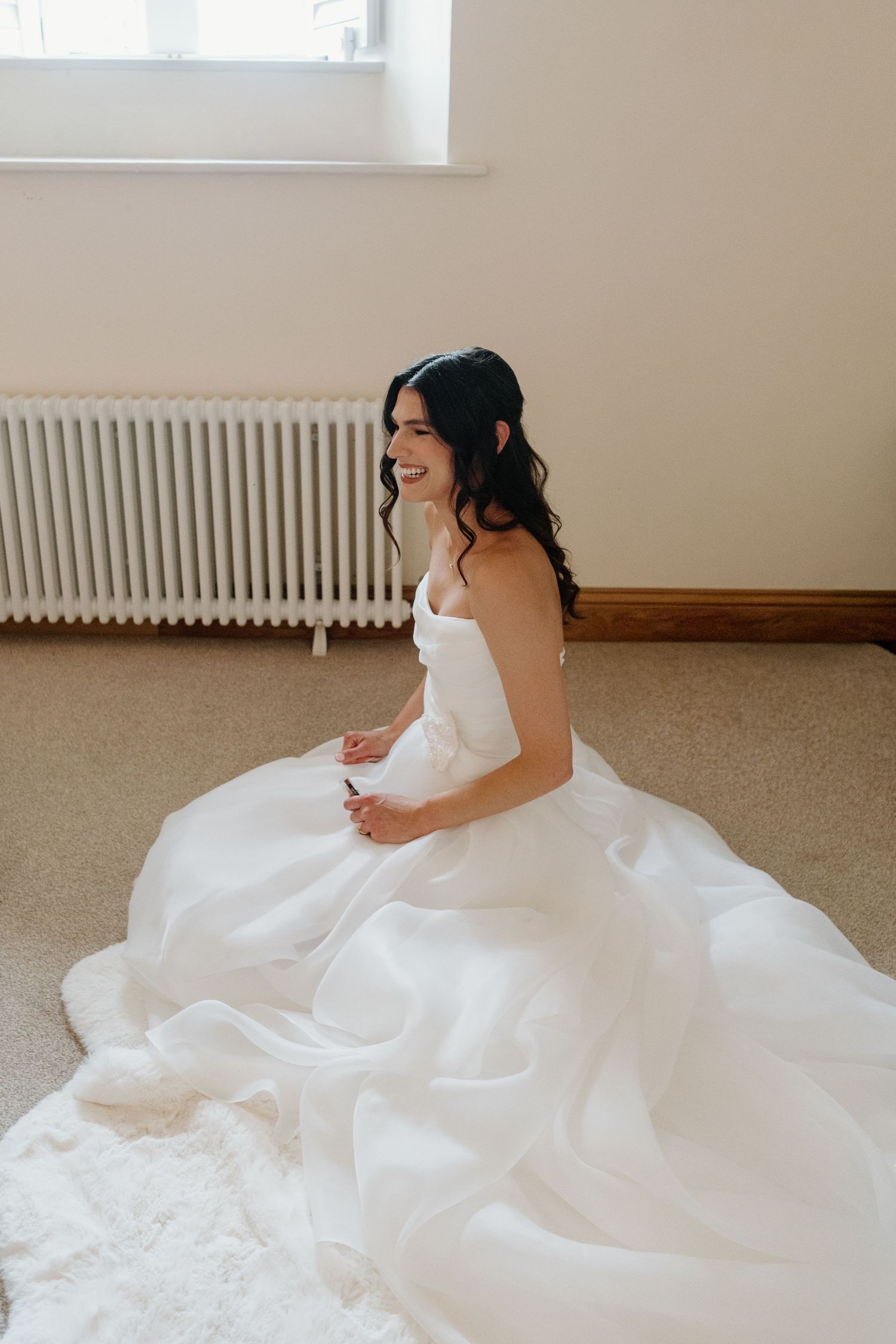Smiling bride in a strapless white ballgown sits on the floor by a bright window.