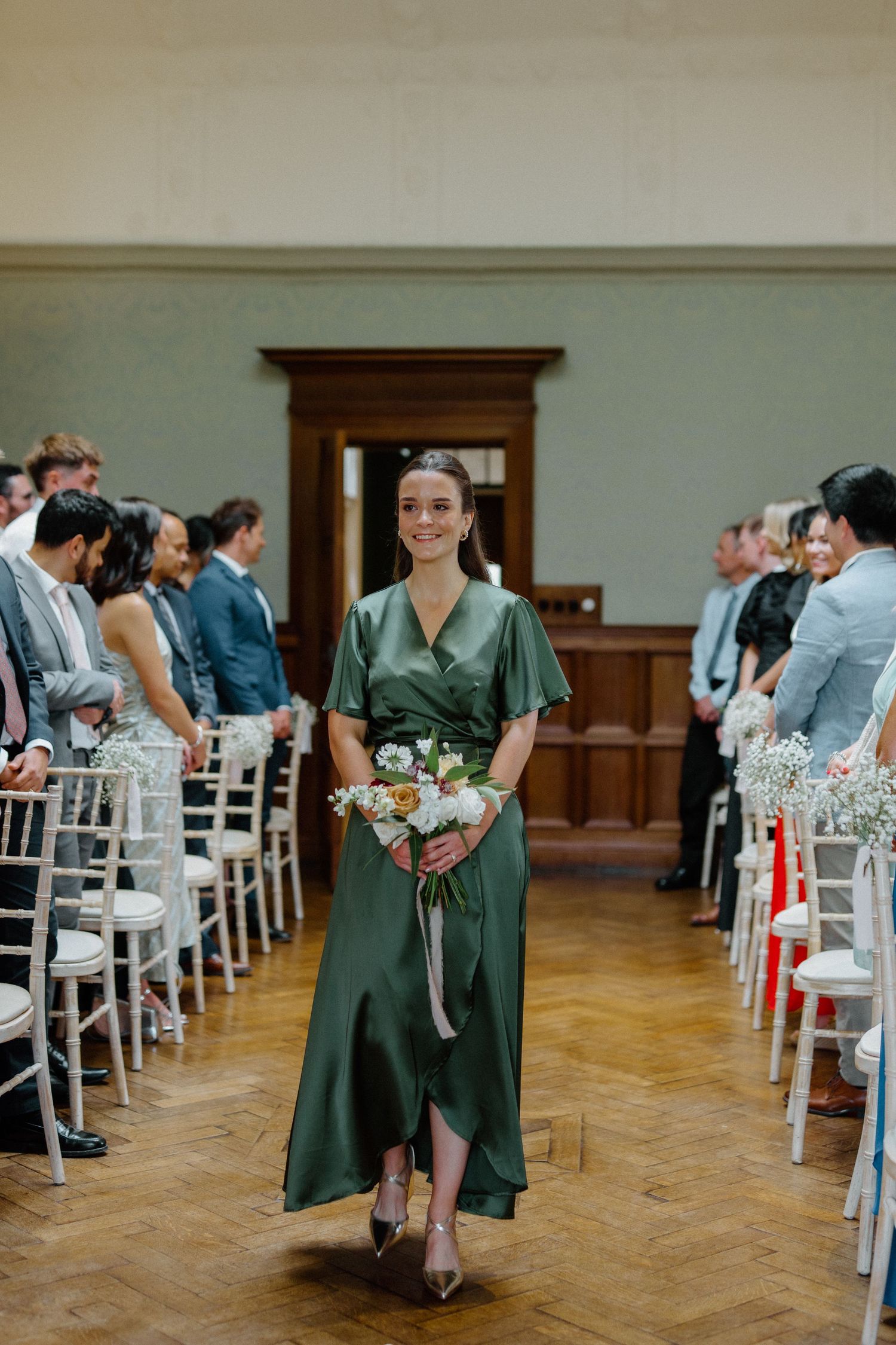 Smiling bridesmaid in emerald green satin dress walking down aisle carrying white bouquet.