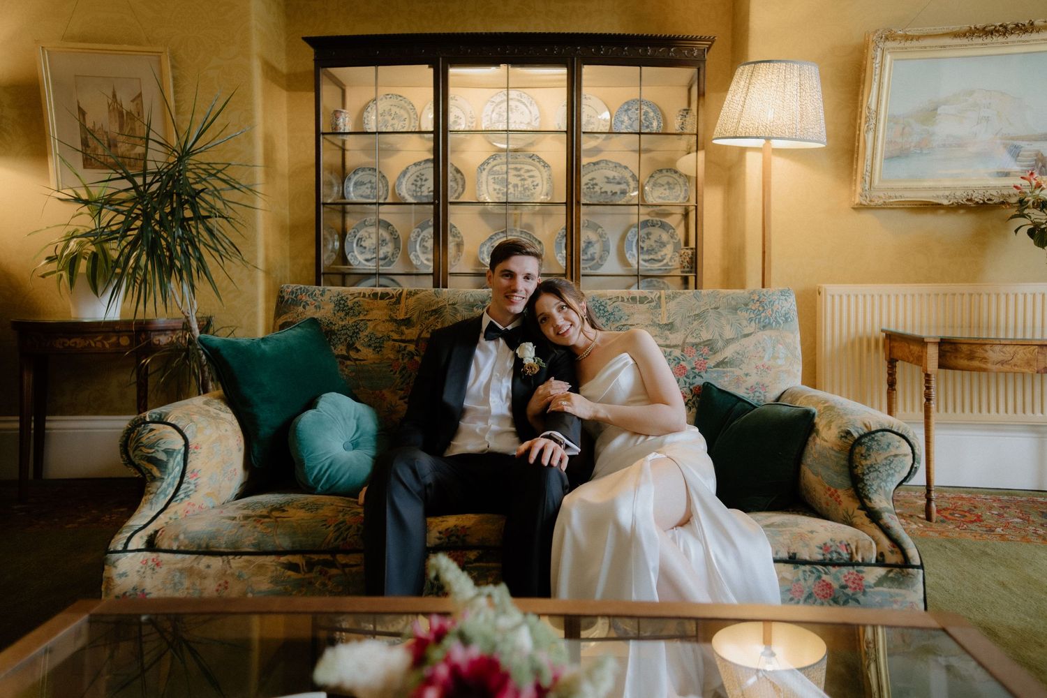 Bride and groom sit together on a vintage floral sofa in an elegantly decorated room with warm lighting.